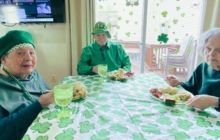 Three people sit at a table with green hats, eating in a bright dining room.