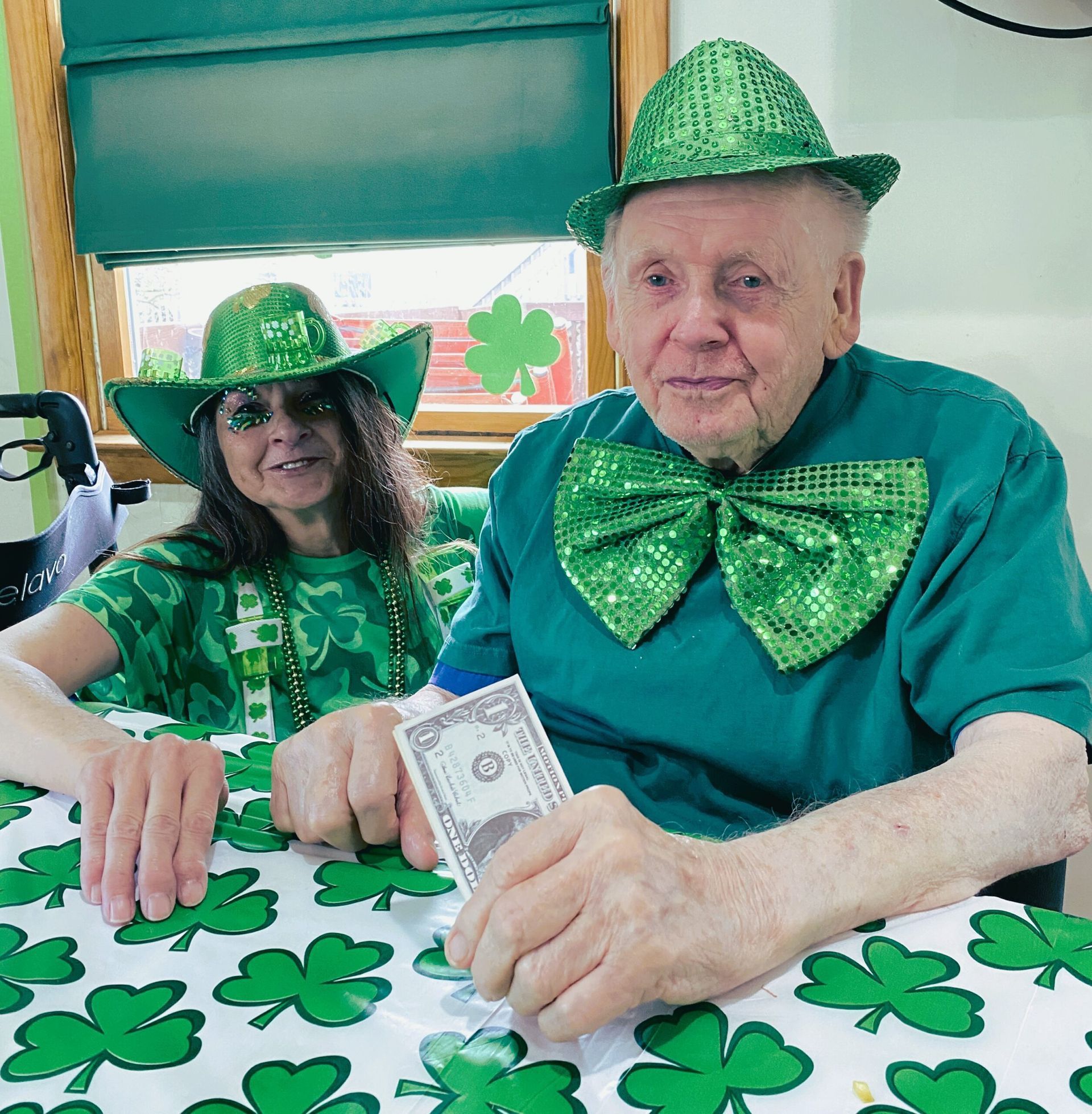 Two people in green St. Patrick’s Day outfits sit at a table decorated with shamrocks, holding money.