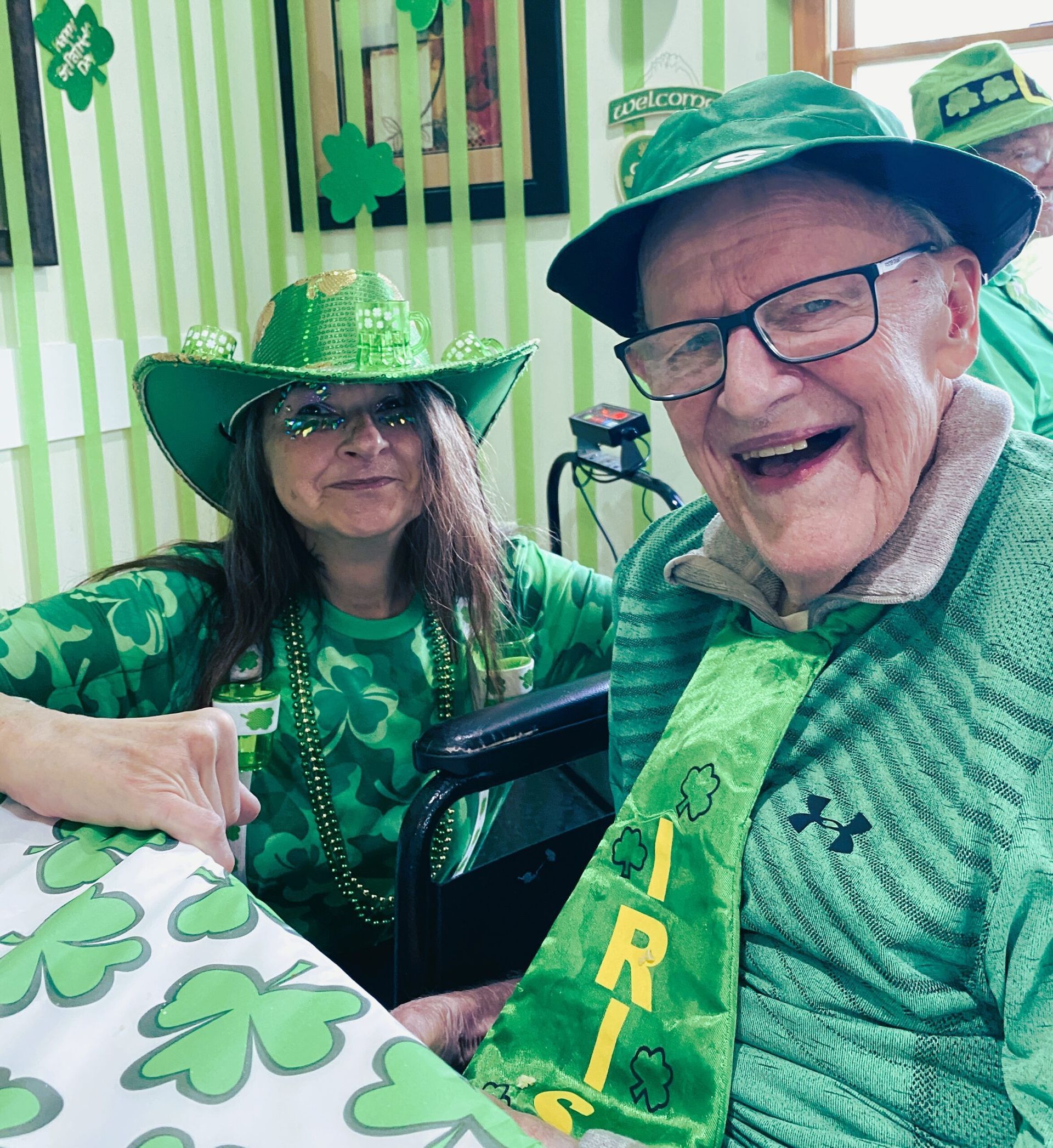 Two people in green St. Patrick’s Day outfits and hats, smiling indoors with shamrock decorations.