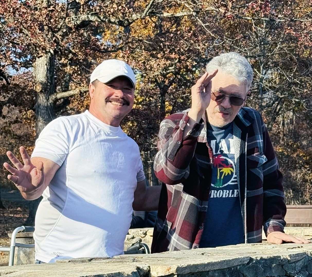 Two people standing outdoors, waving and smiling by a stone wall in autumn trees