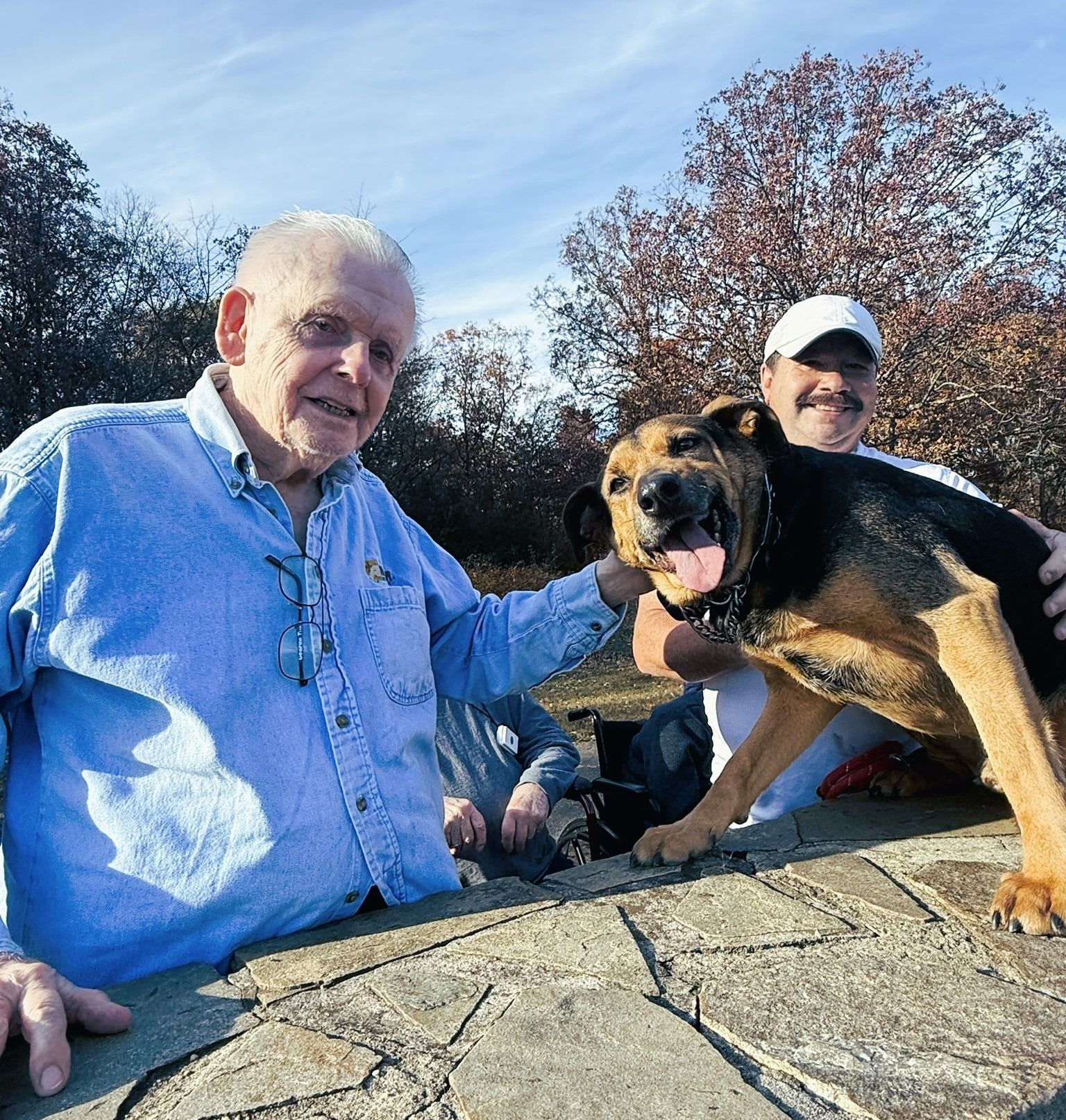 Two people smile beside a black-and-tan dog outdoors on a sunny day