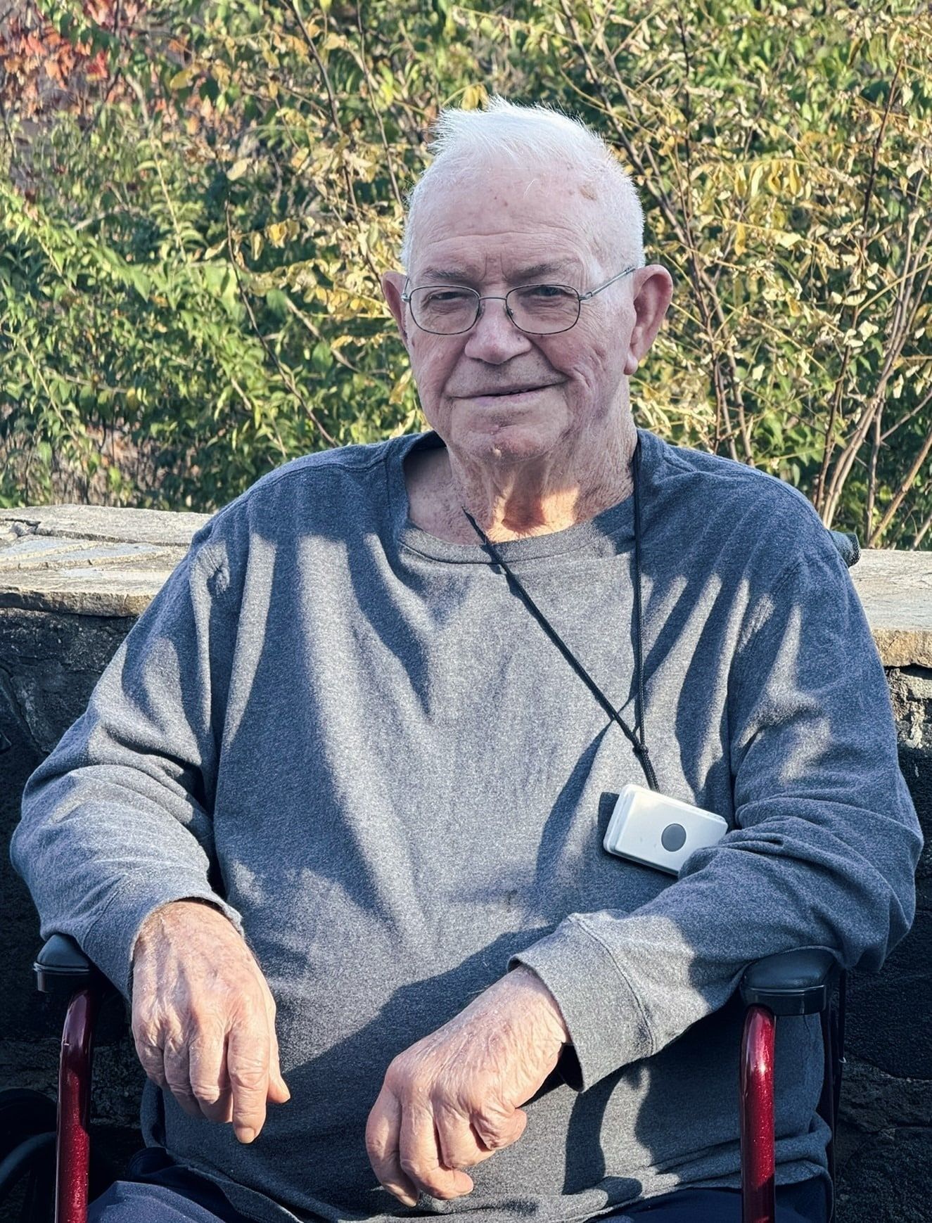 Smiling man in a gray shirt sitting outdoors in a chair, with green foliage behind him.
