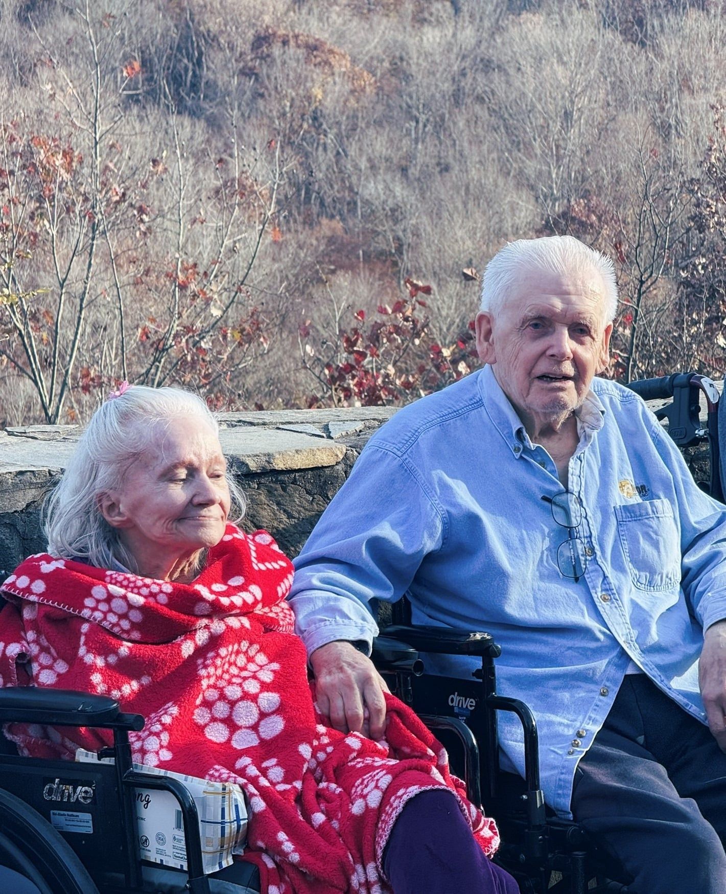 Two people seated outdoors, one wrapped in a red patterned blanket, in front of a wooded hillside.