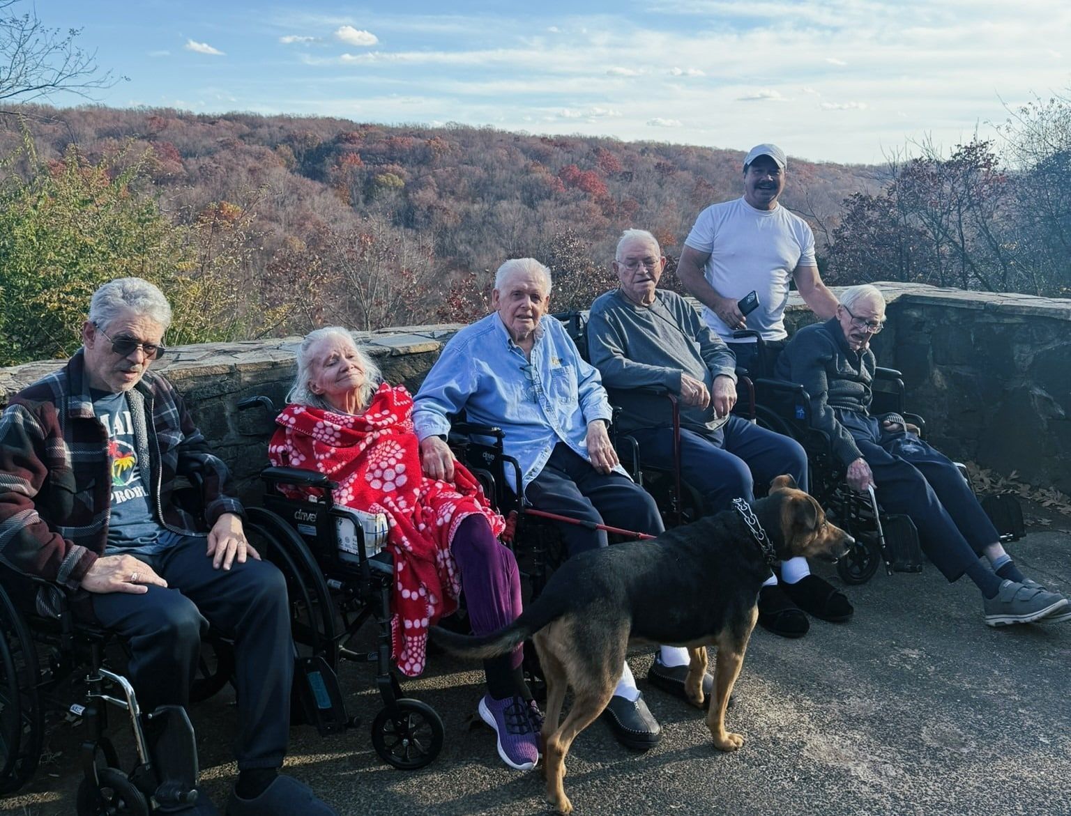 Five people seated outdoors on a scenic overlook, with two dogs in front and hills in the background.