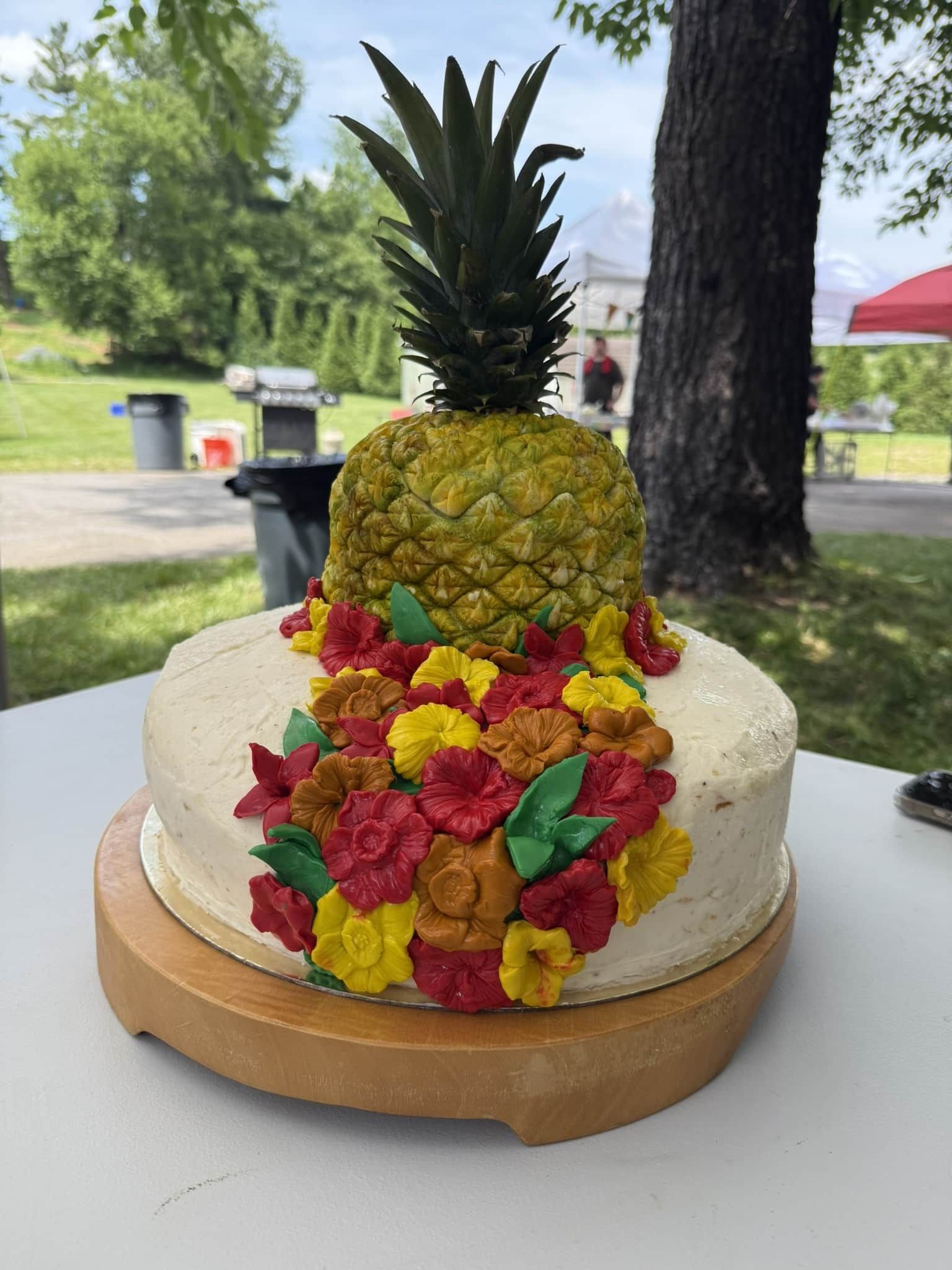 Pineapple-shaped cake with red and yellow flowers on a wooden stand at an outdoor picnic table