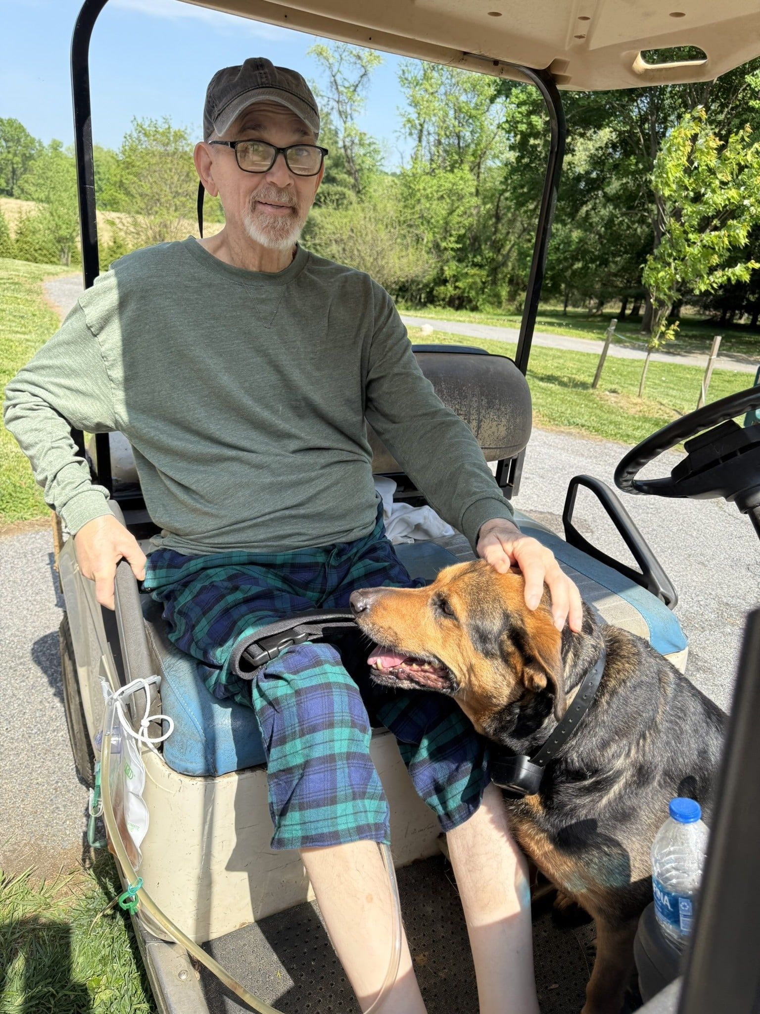 Man seated in a golf cart with a dog on his lap, wearing a cap and plaid shorts on a sunny day.