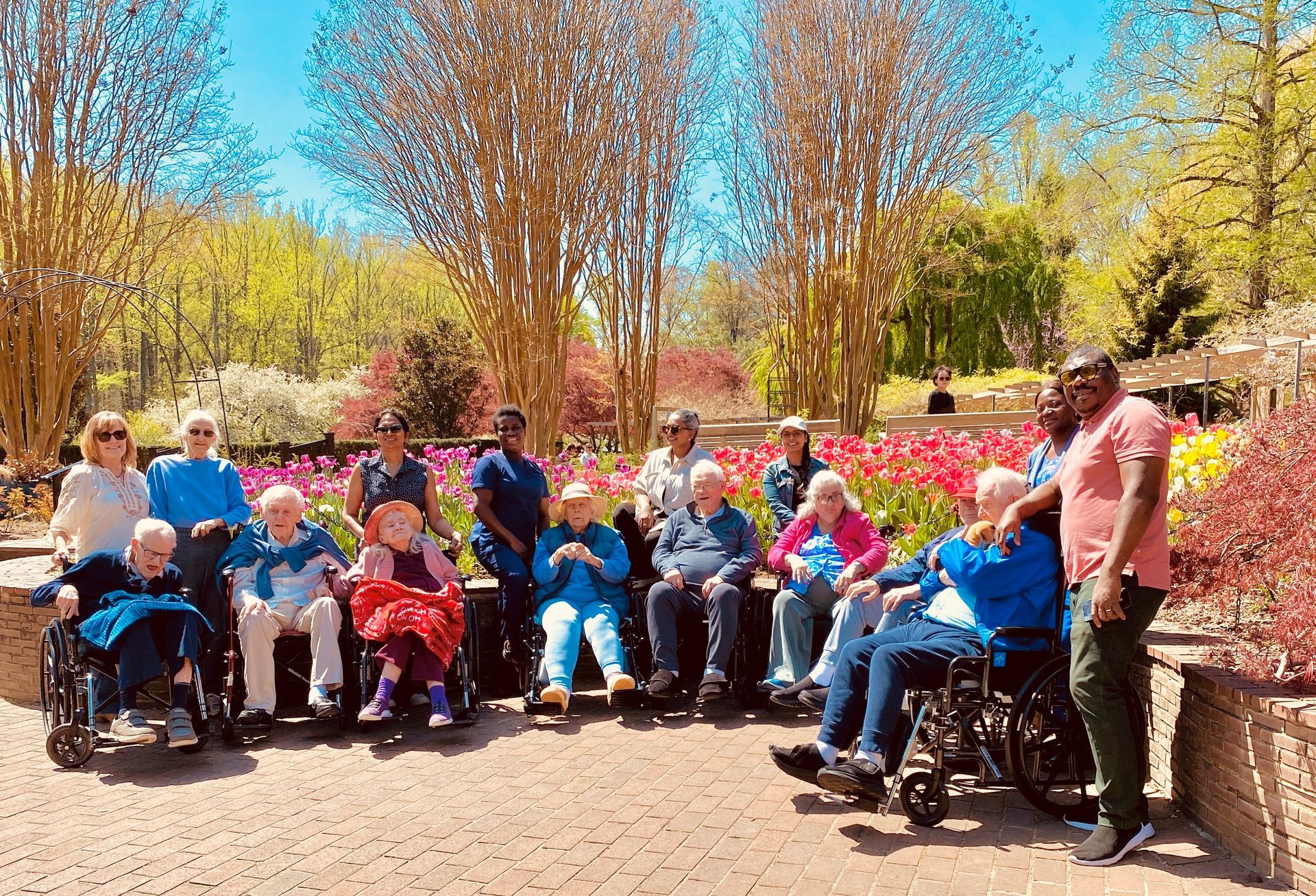Group of people seated outdoors in wheelchairs and on benches in a sunny garden with autumn trees.