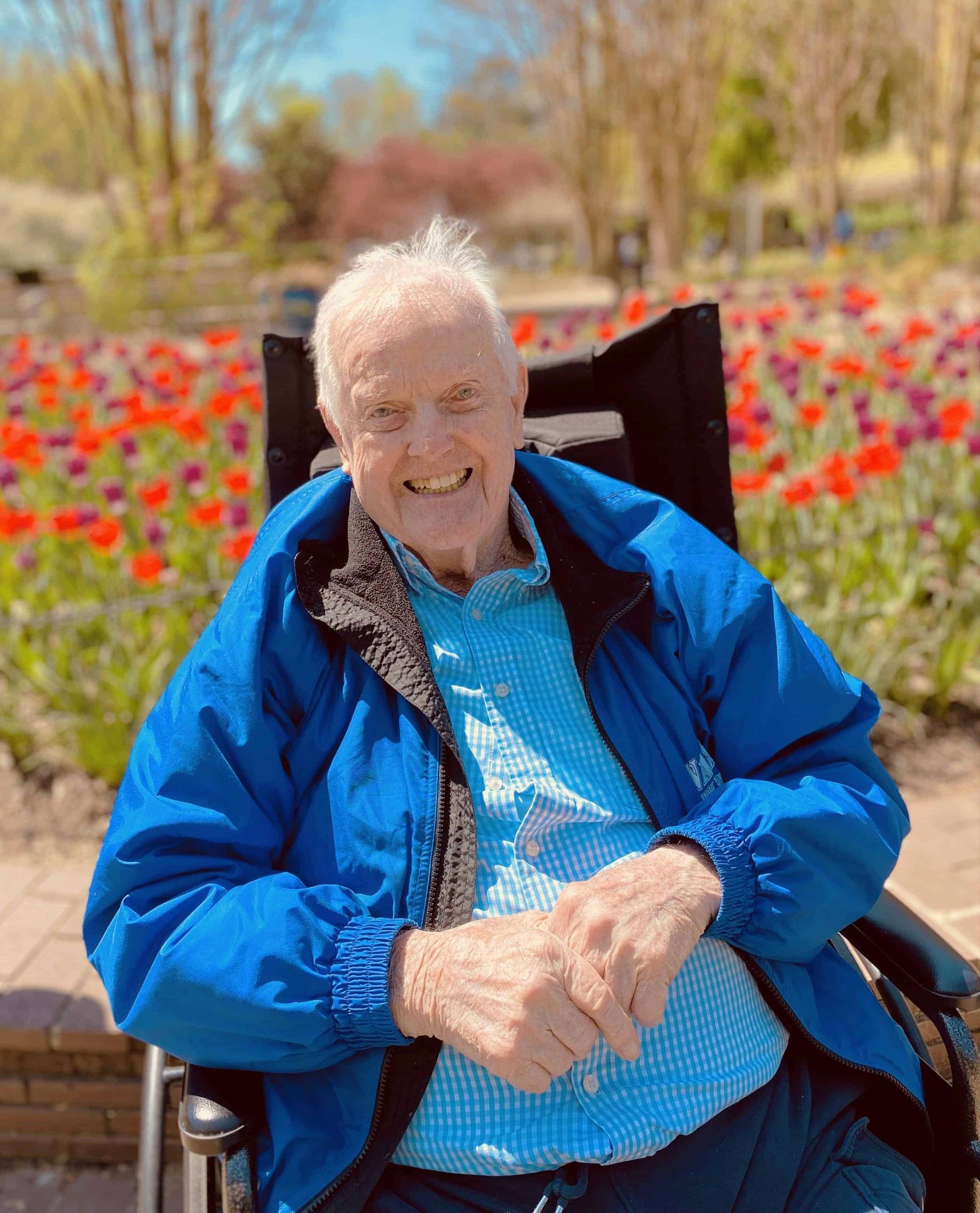 Smiling person in a blue jacket sitting outdoors among red flowers and trees