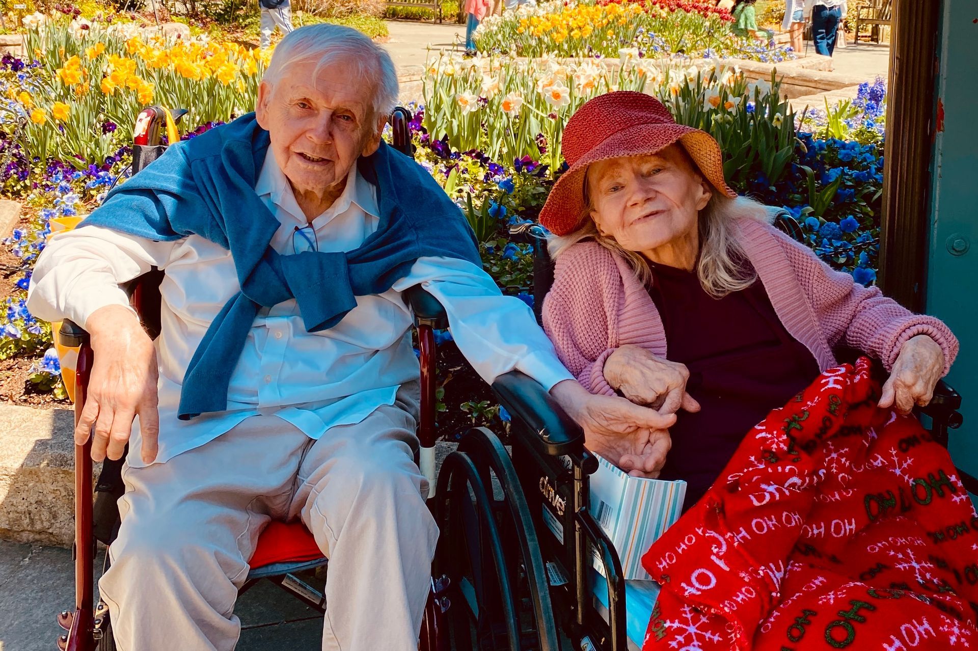 Two elderly people sitting together on a bench in a colorful flower garden, smiling and holding hands