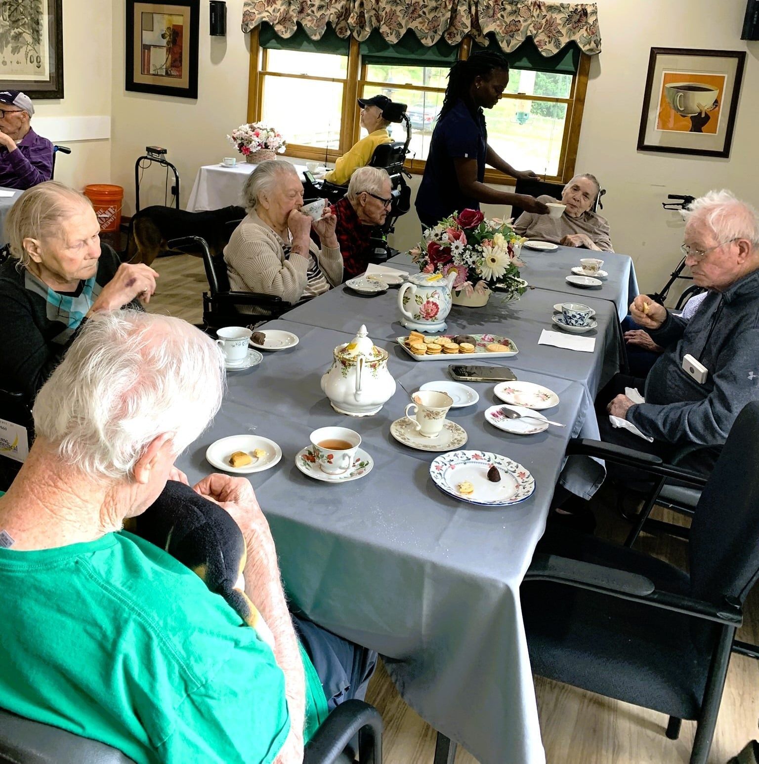 People seated around a tea table in a cozy living room, chatting over cups and plates of desserts.