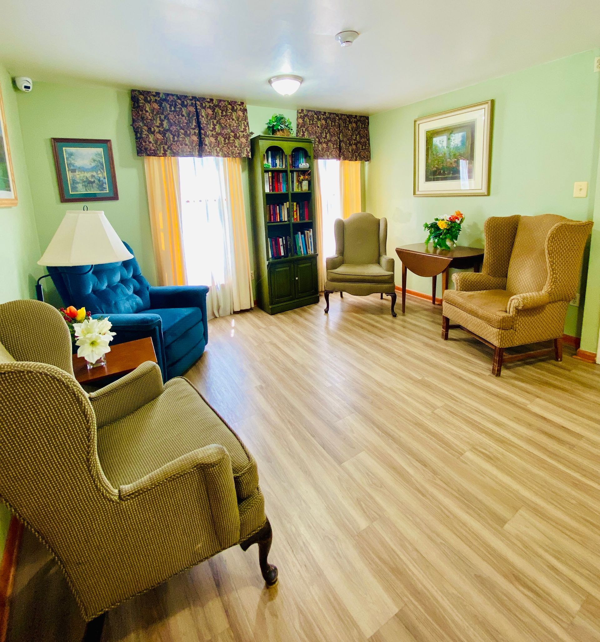 Cozy lounge with wicker chairs, blue armchair, bookshelf, and patterned window curtains in soft green walls.
