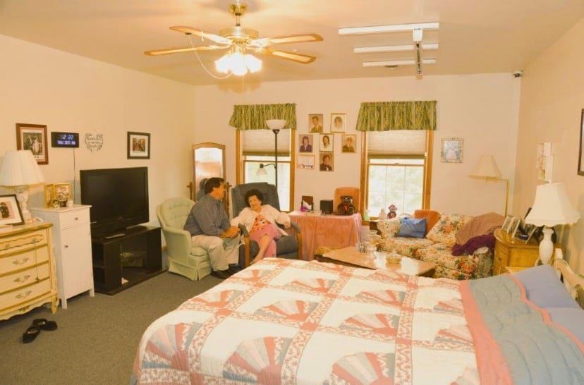 Bedroom with a quilted bed, dresser, TV, and two people sitting near the window in a cozy, cluttered room