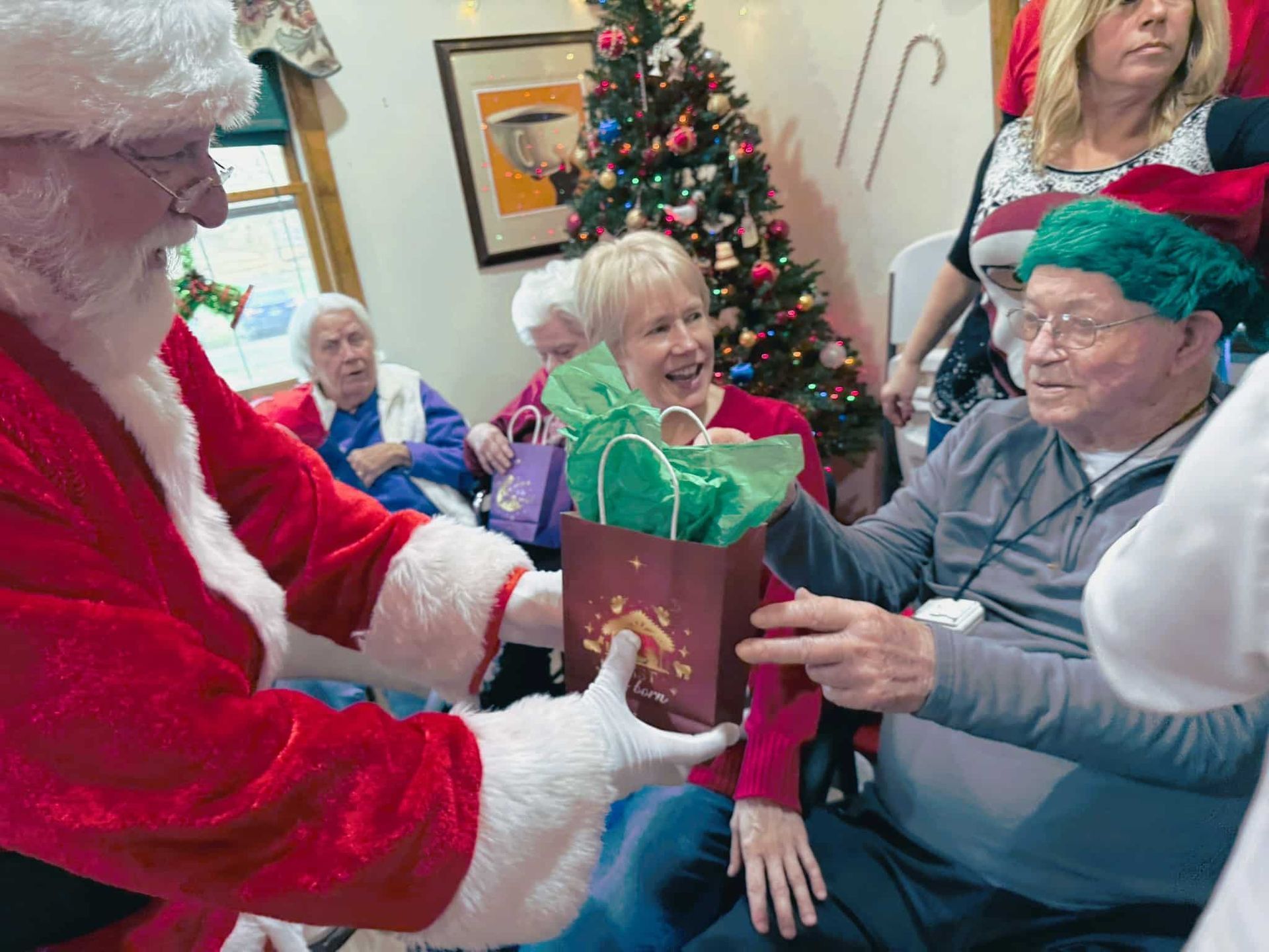 Santa handing a gift bag to a smiling seated man at a Christmas gathering
