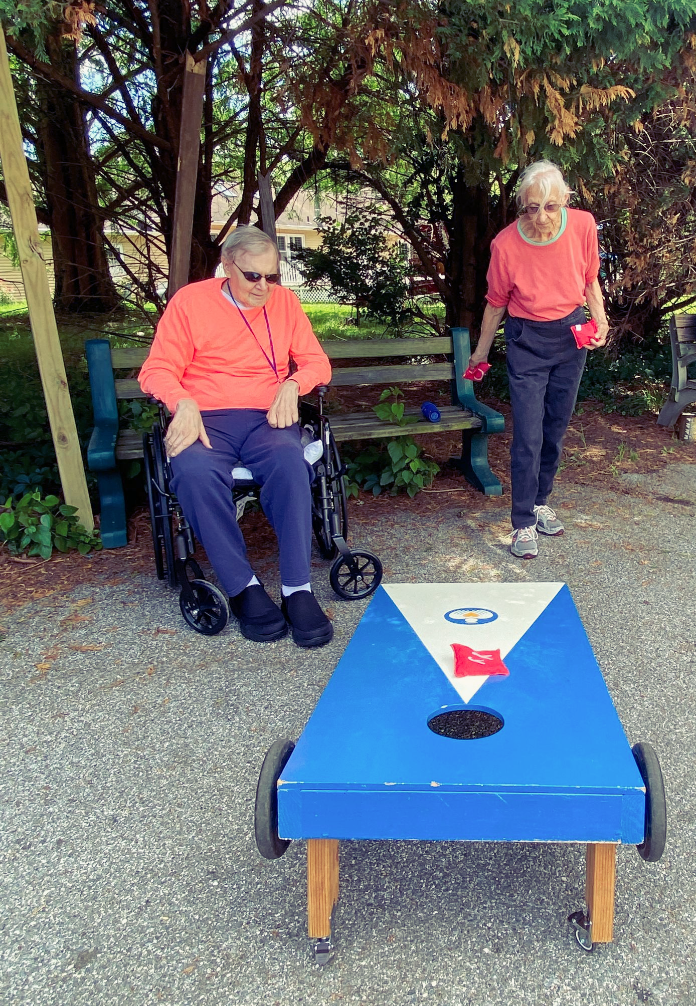 Two people playing cornhole outdoors, with a blue board and beanbags on gravel under trees.