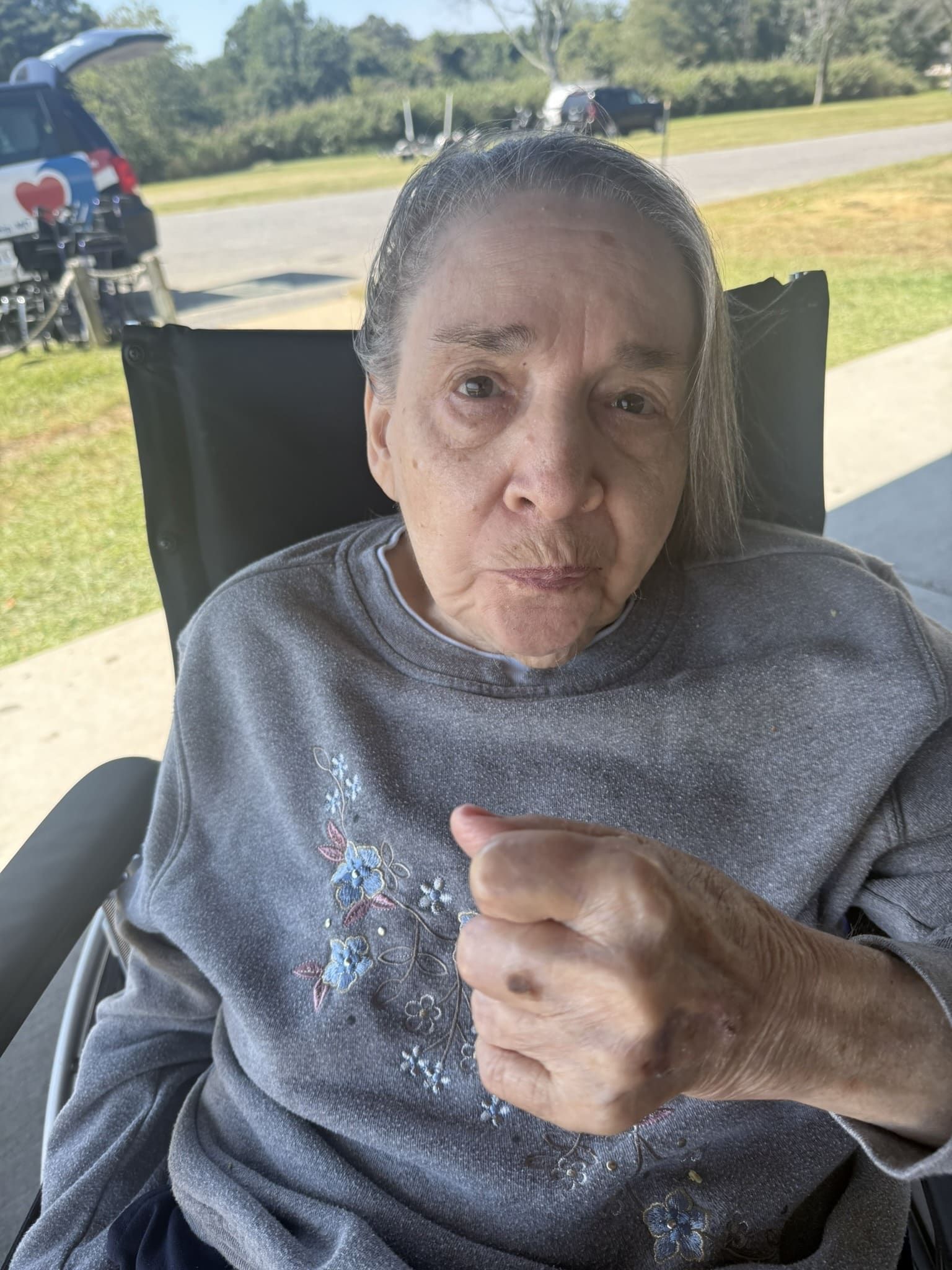 Person seated outdoors in a chair, wearing a gray sweater and giving a thumbs-up.