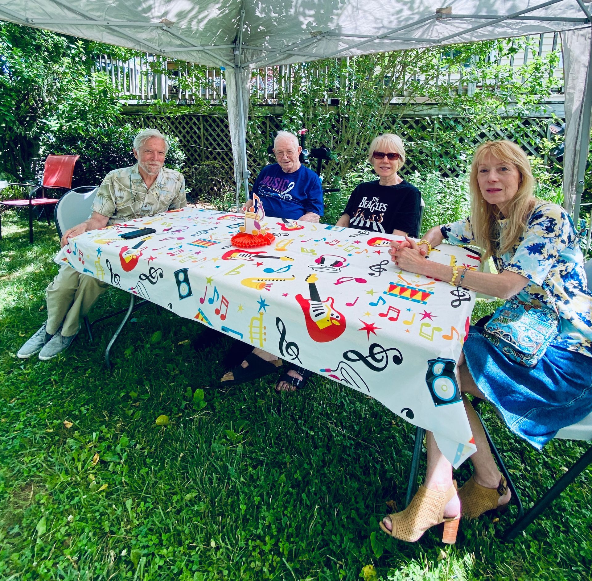 Four people sitting at a decorated outdoor table under a tent in a green garden, smiling.