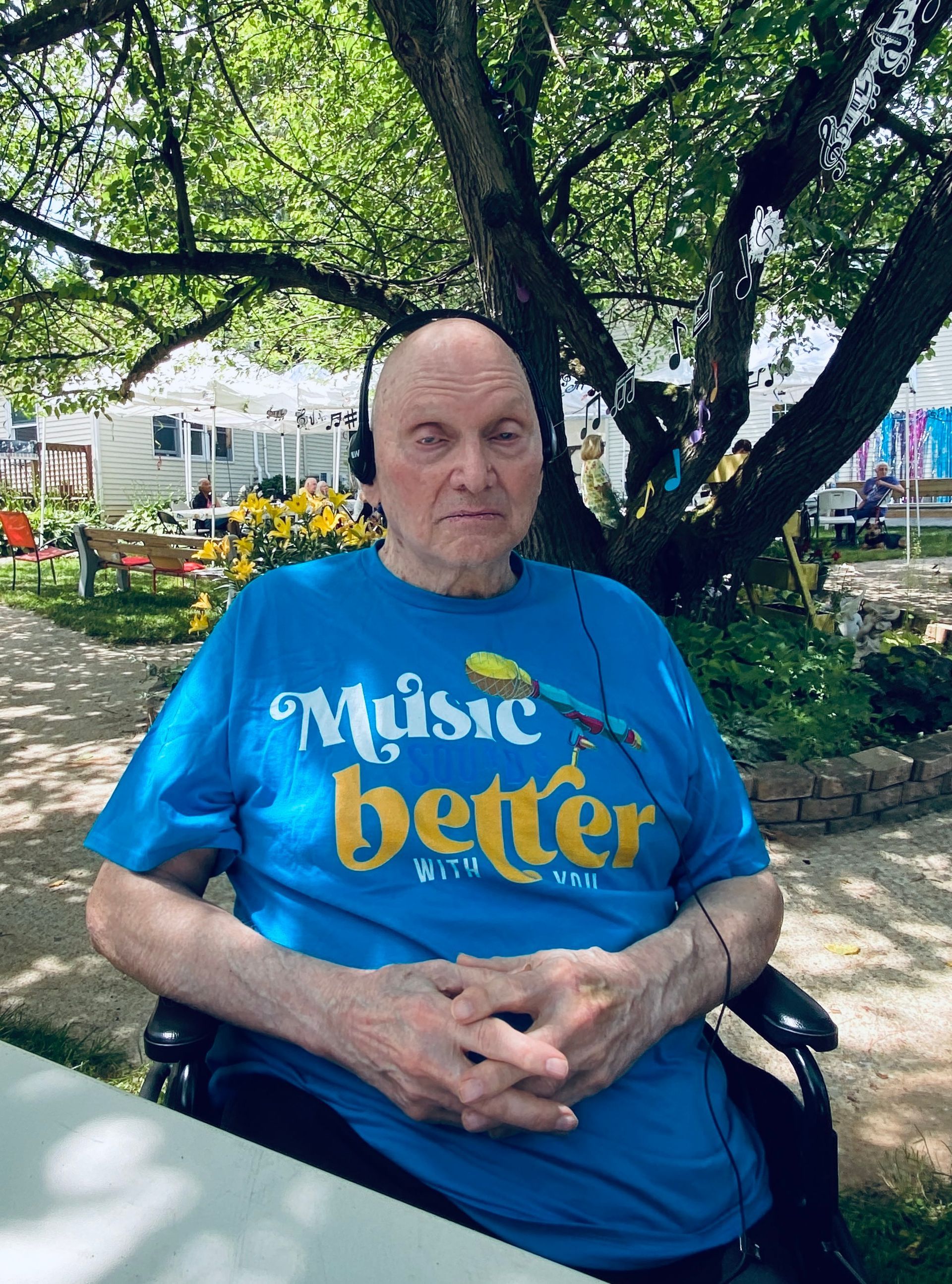 Person seated outdoors under a tree, wearing a blue “Music better” T-shirt.