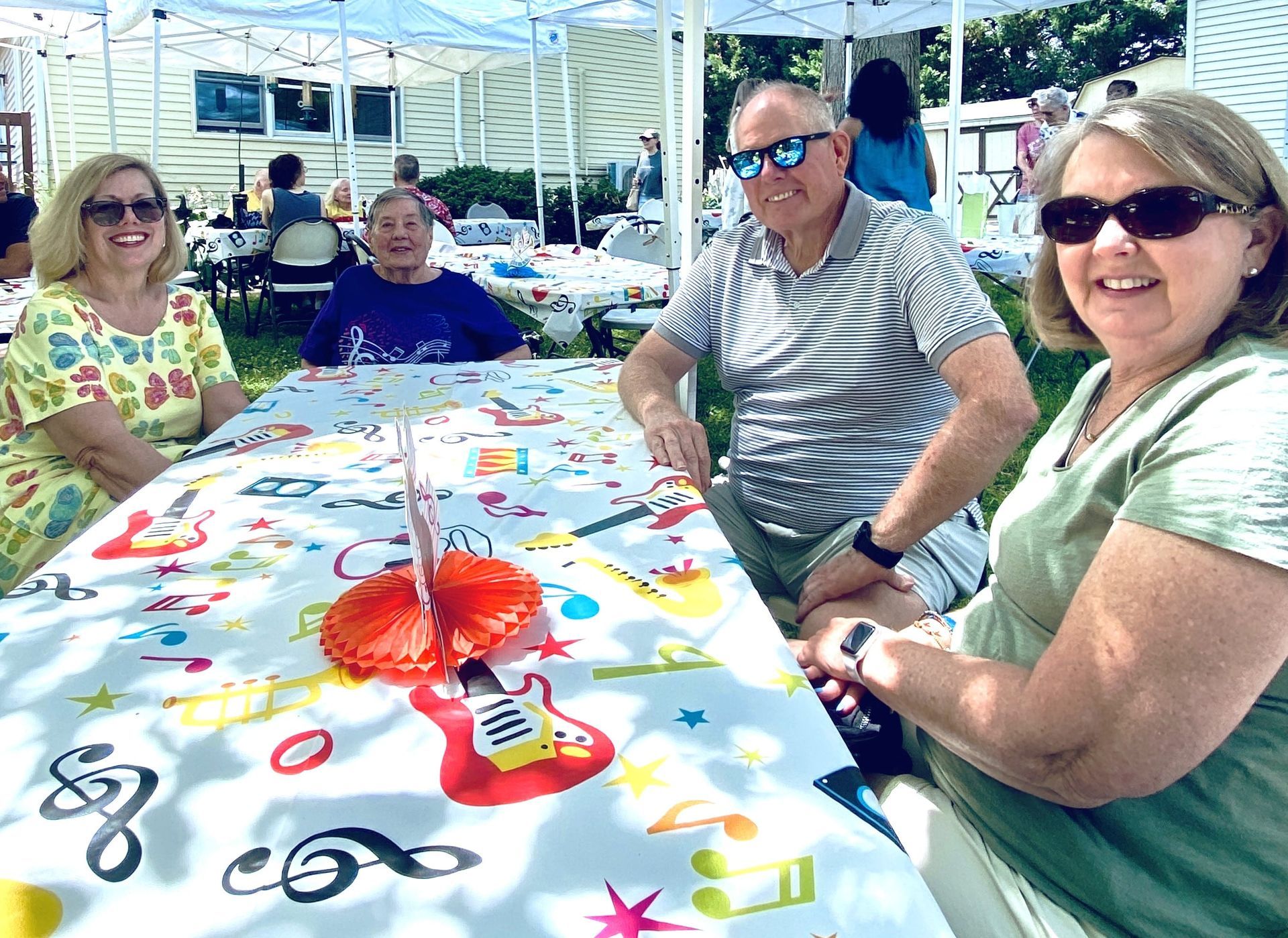People sitting around a colorful tablecloth at an outdoor picnic, smiling in a shaded pavilion.