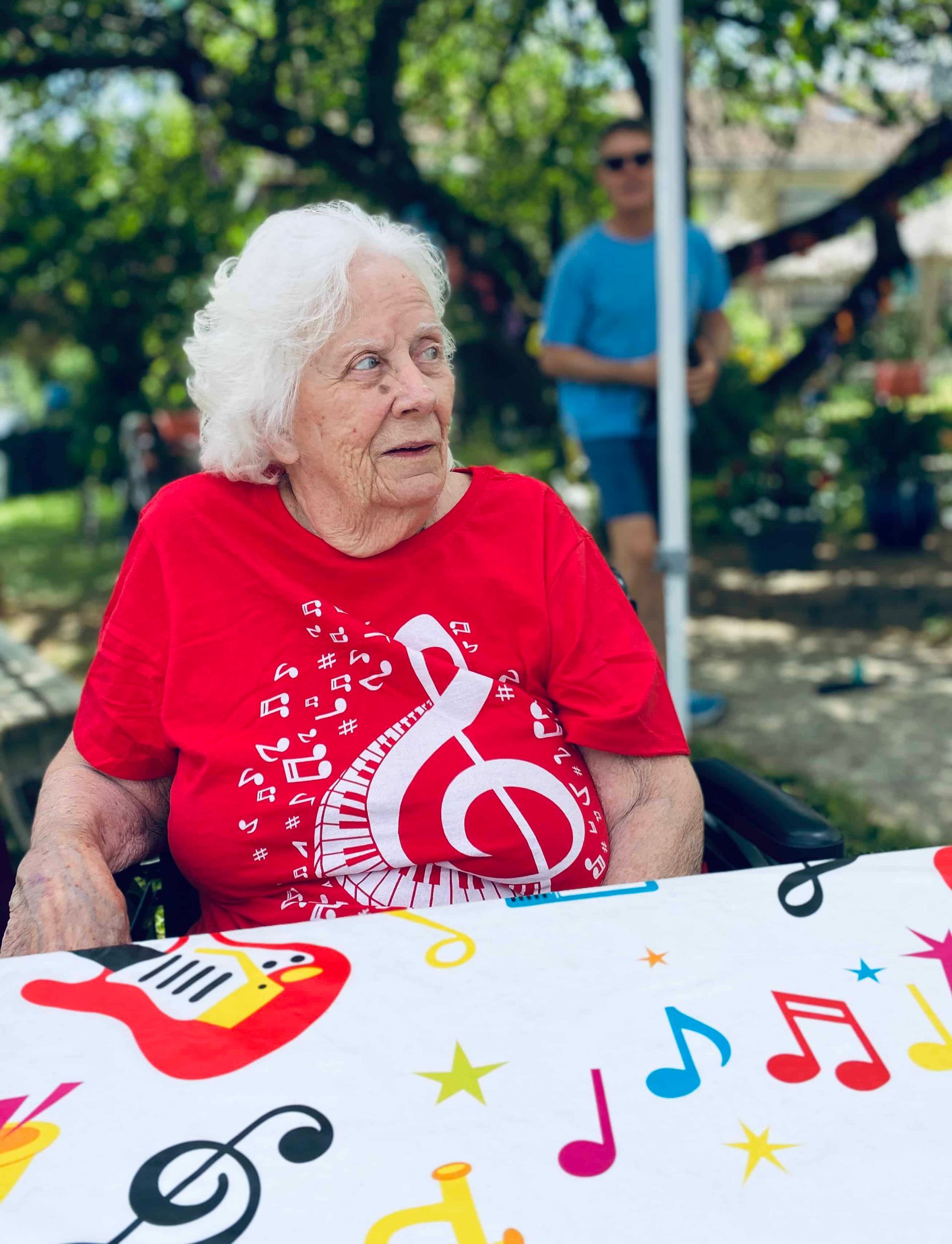 Elderly woman in a red music-themed shirt seated outdoors at a decorated table in a park