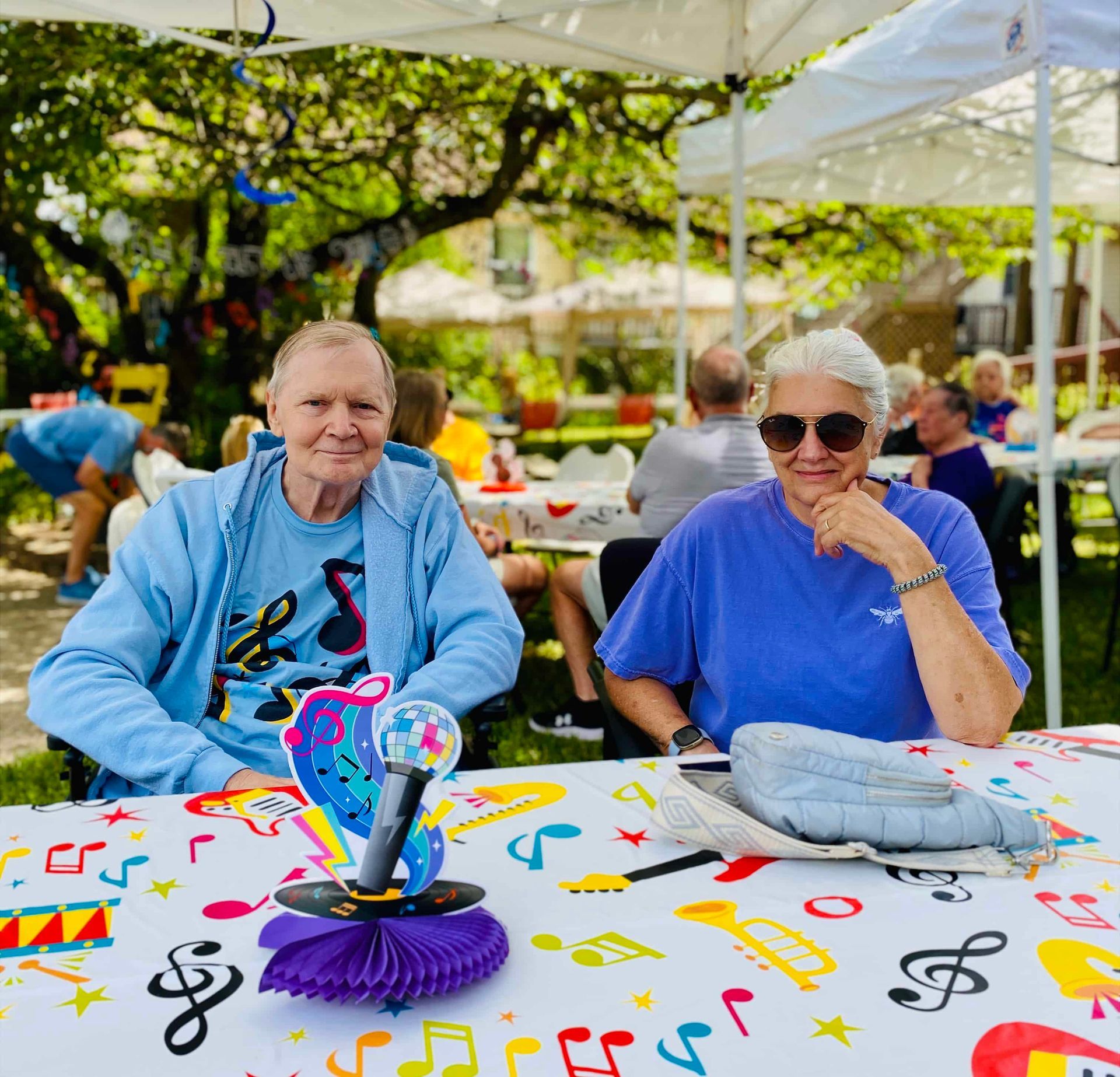Two people seated at a colorful outdoor table under a canopy, with a trophy centerpiece.