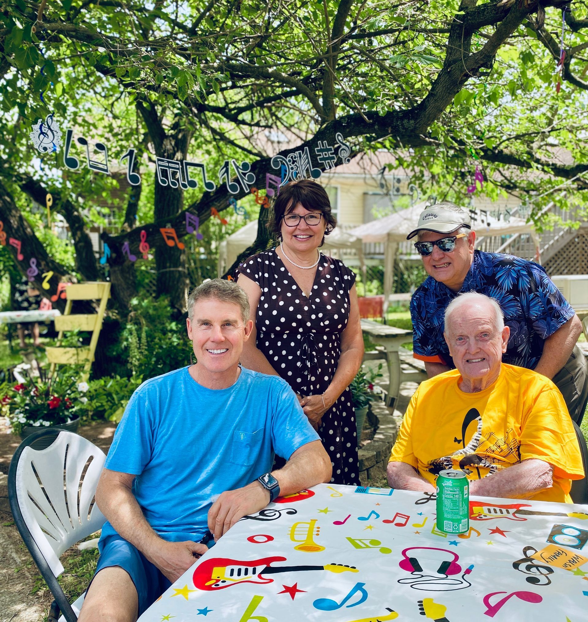 Four people smiling at an outdoor table under a leafy tree at a backyard gathering