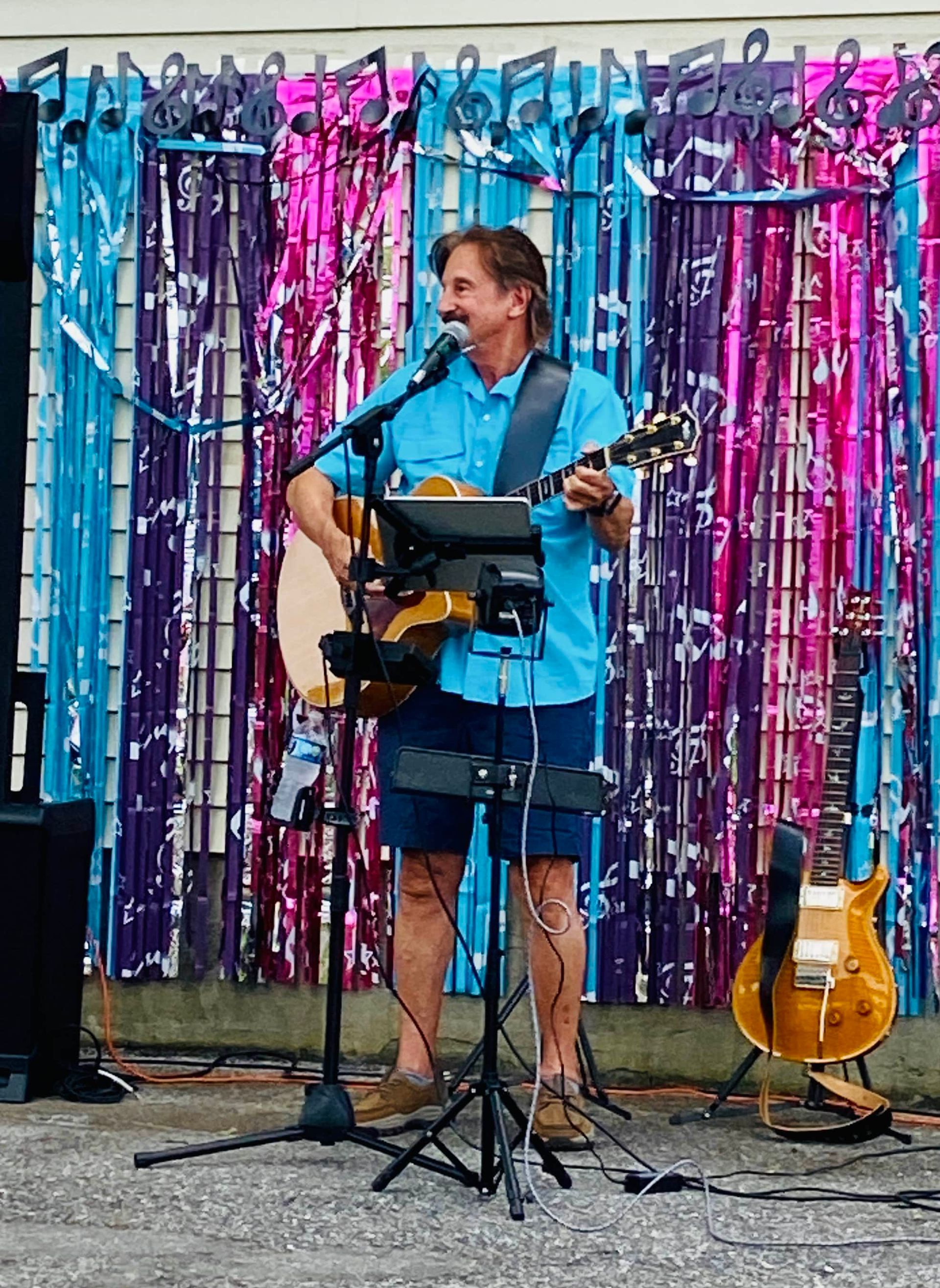 Man in blue shirt singing and playing acoustic guitar on stage with colorful tinsel backdrop