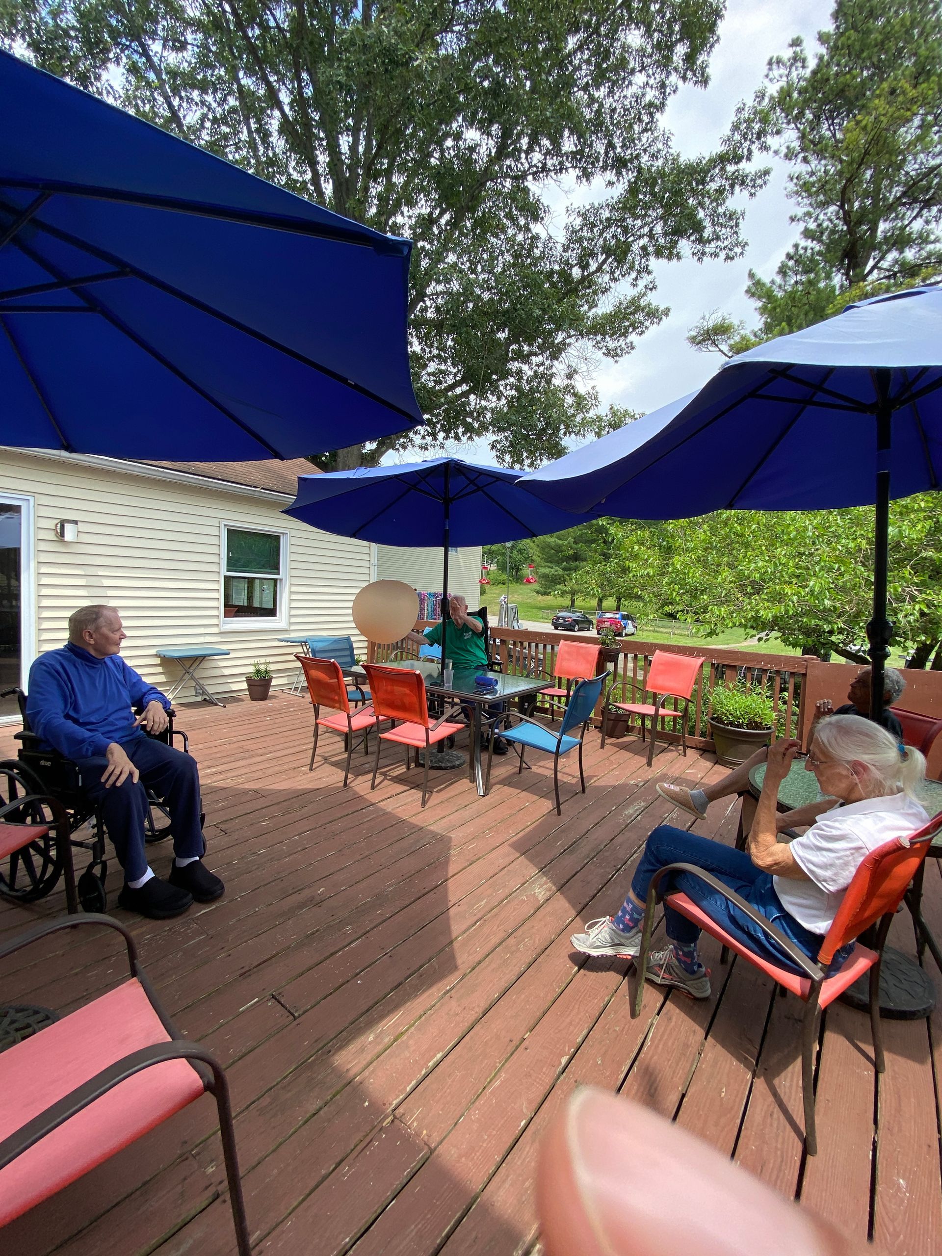 People sitting on a wooden deck under large blue umbrellas, surrounded by trees and a house.