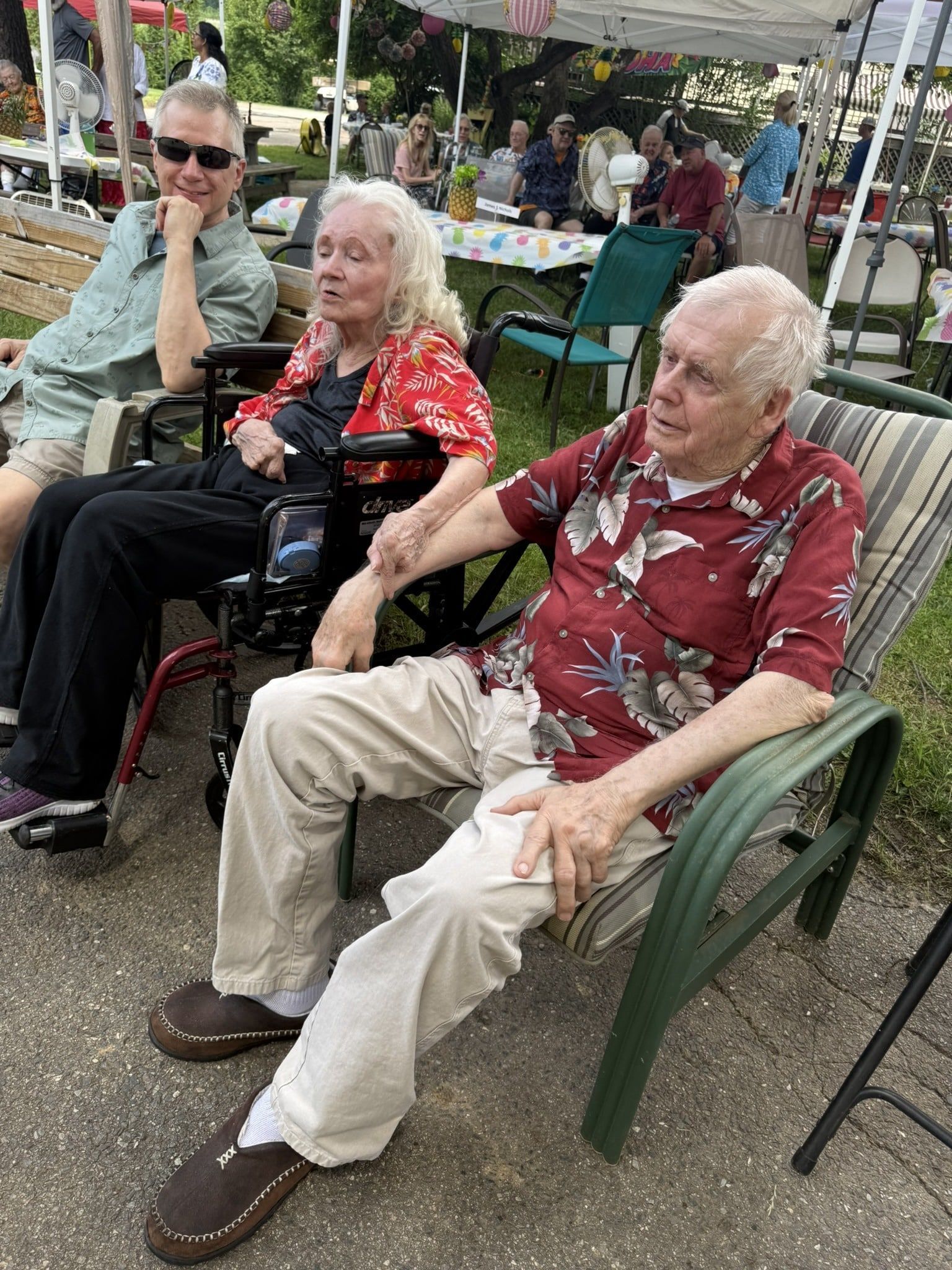 Three people seated outdoors at a community gathering, with tables and flowers in the background.