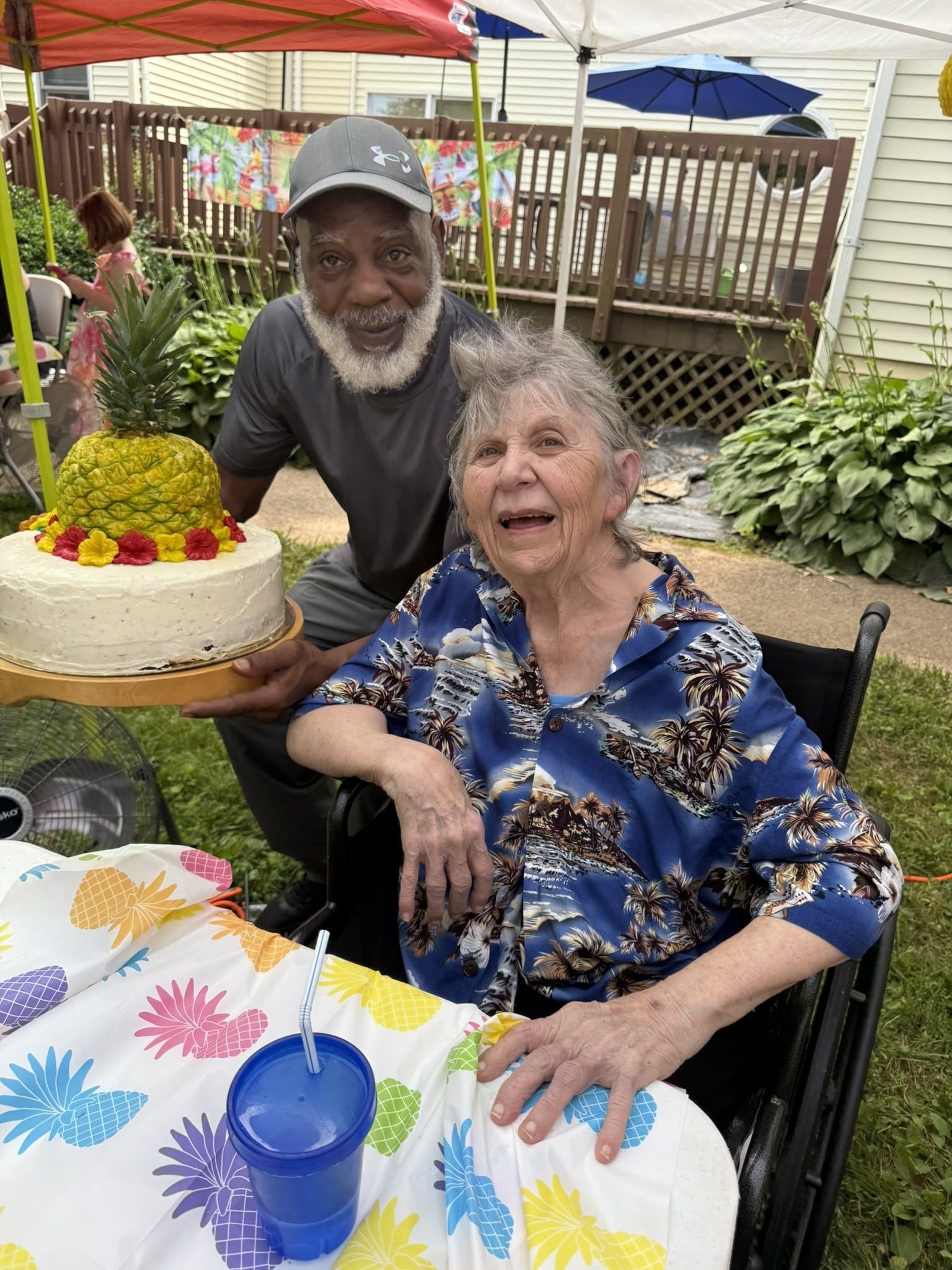 Two people smiling at a decorated birthday cake outdoors, seated at a table with a blue cup and colorful tablecloth