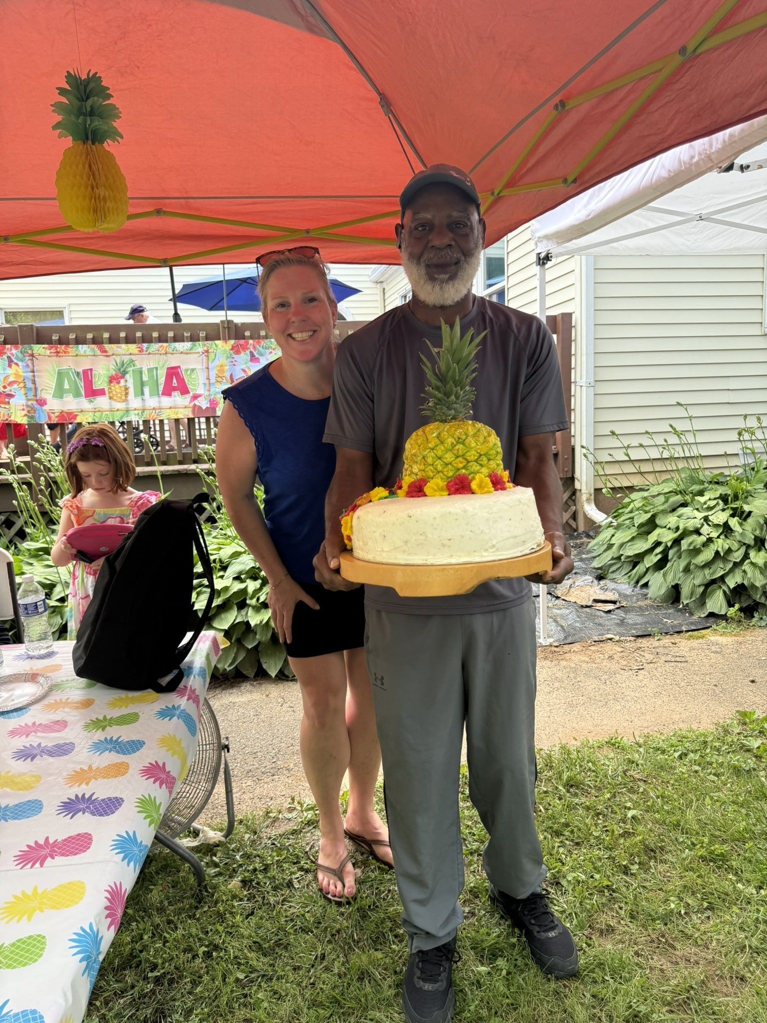 Two people holding a decorated cake under a pink canopy at an outdoor event.