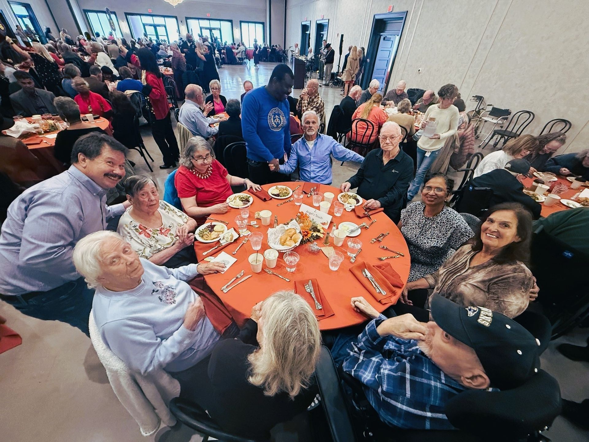 People seated around orange-clothed tables at a lively indoor gathering, chatting and eating.