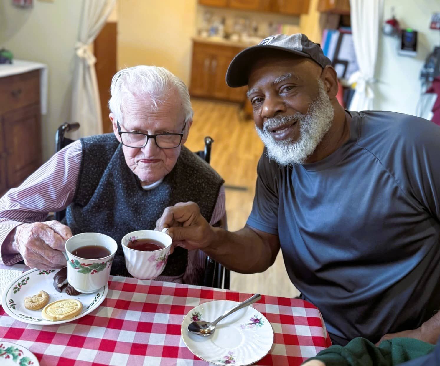 Two people smiling at a red-checkered table, raising mugs in a cozy kitchen