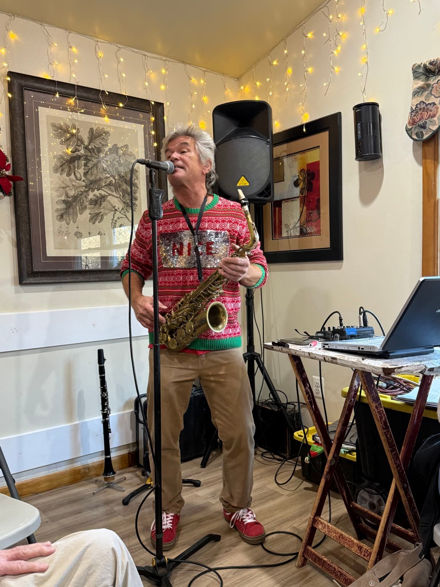 Man playing saxophone in a cozy room with string lights, speaker, mic stand, and laptop.