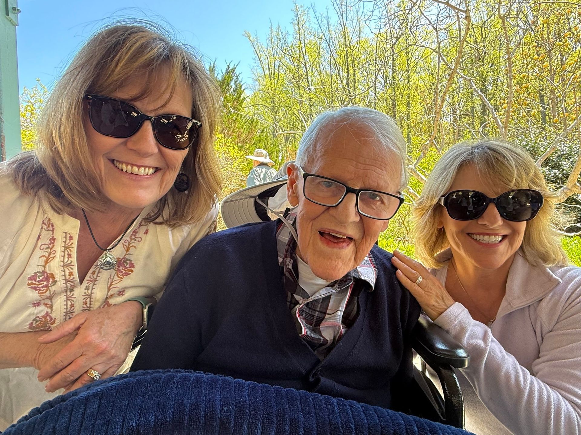 Three smiling people outdoors in sunlight, two leaning in beside an older man in glasses and a wheelchair