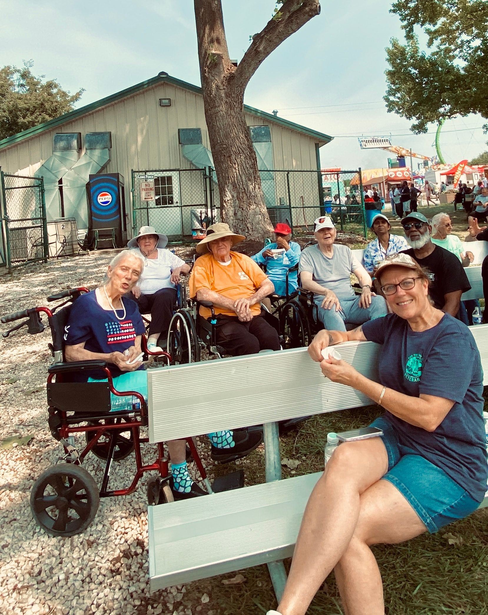 People at an outdoor gathering sit on benches near a tree and a building.