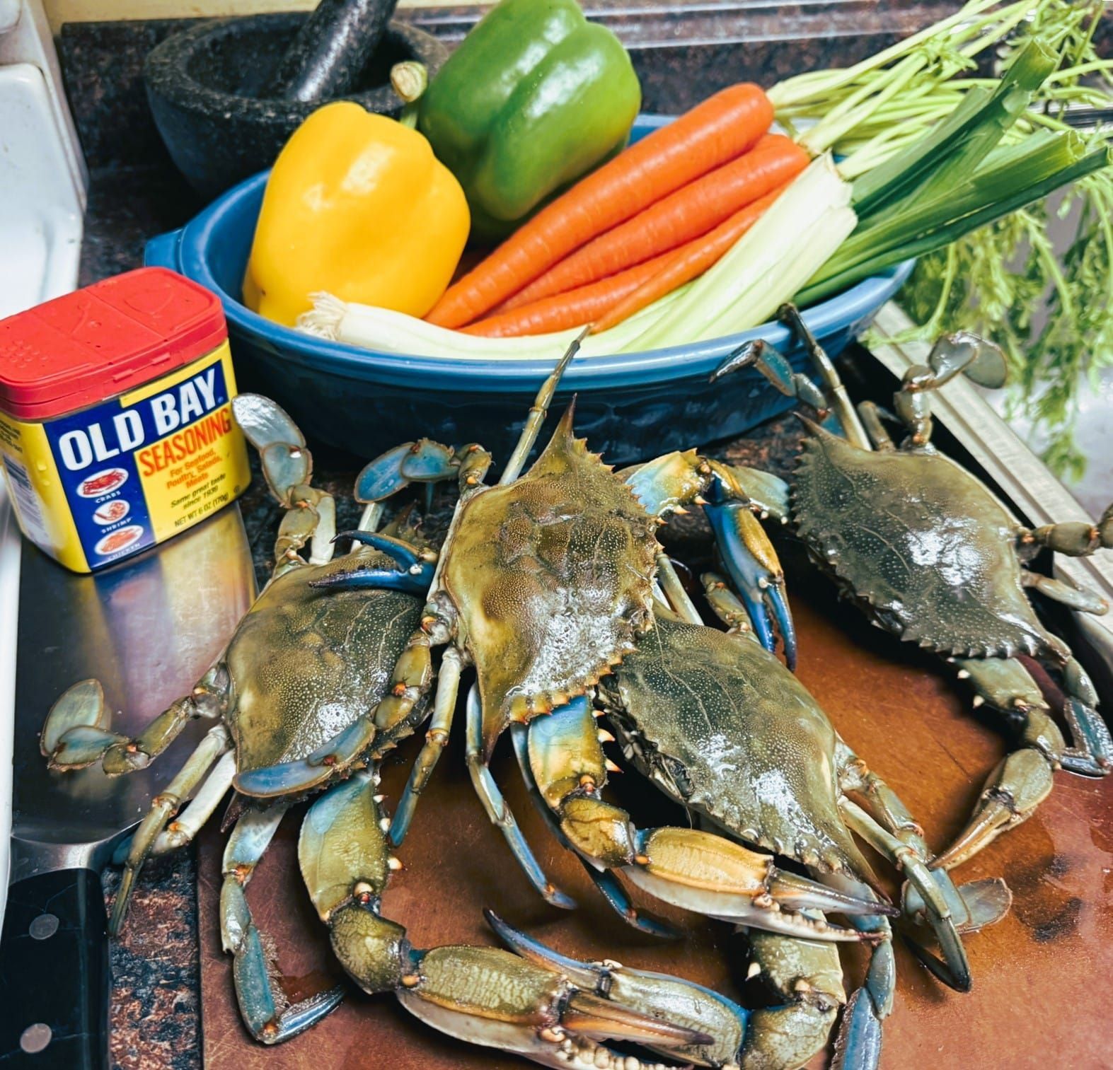 Crabs on a cutting board beside Old Bay seasoning and a bowl of colorful vegetables and herbs