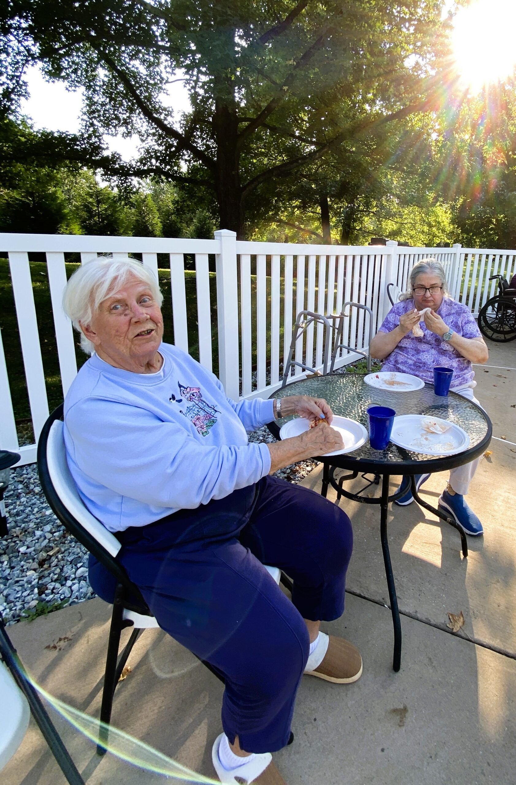 Two women seated outdoors at a table with drinks, talking beside a white fence in bright sunlight