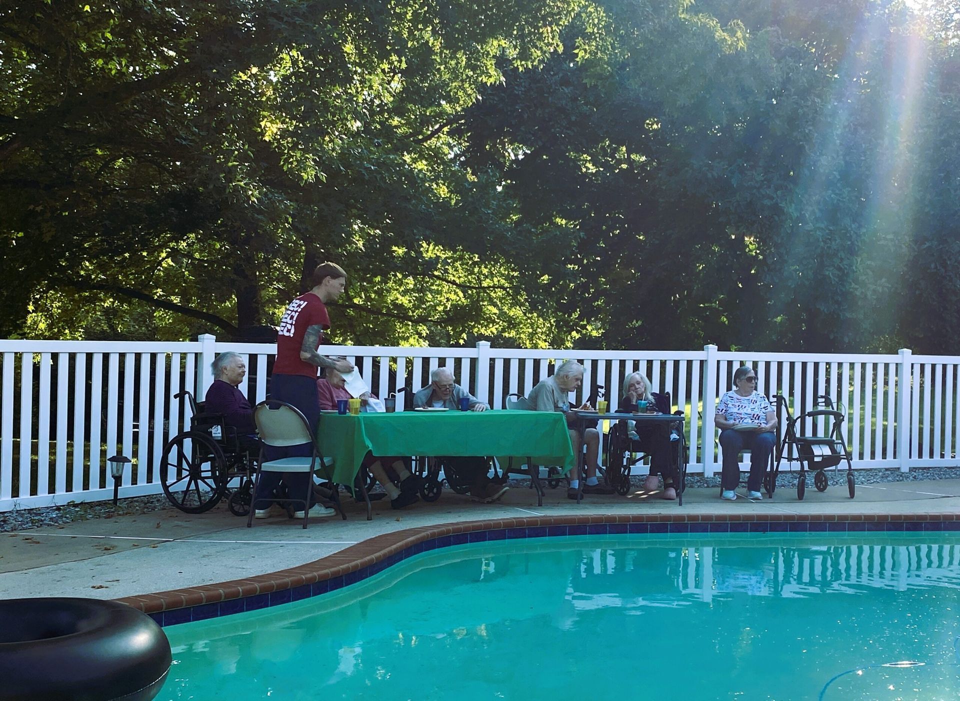 Poolside gathering under trees, with people seated by a white fence beside a turquoise swimming pool.