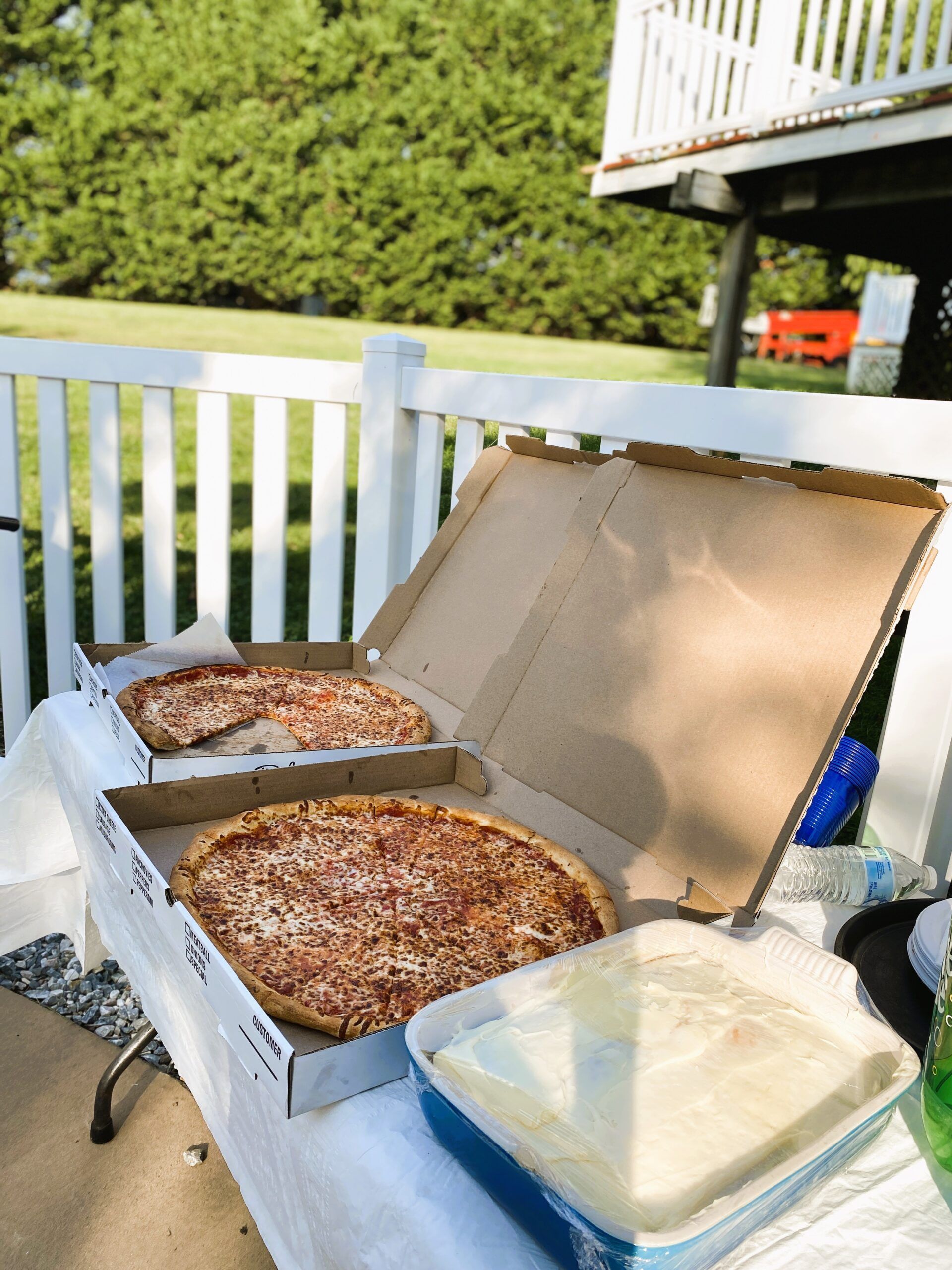Open pizza boxes and a tray of frosted cake on an outdoor table by a white railing.