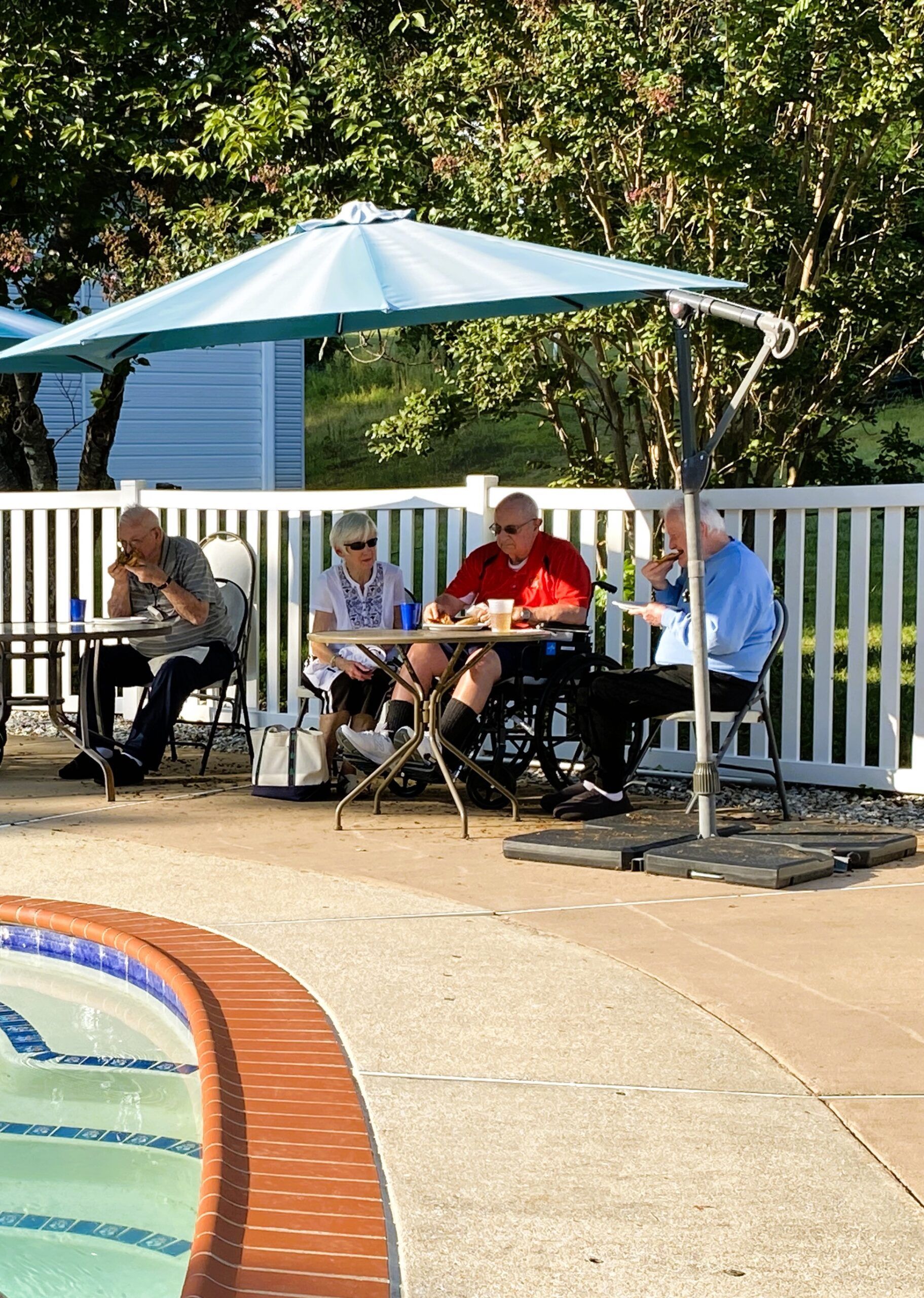 People seated at tables under a white patio umbrella beside a pool and fence