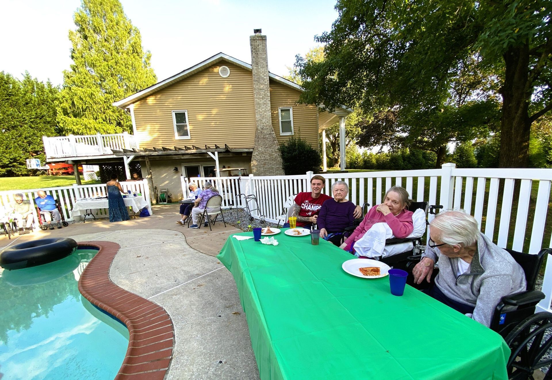 People seated at a green table beside a backyard pool, with a house and patio in the background