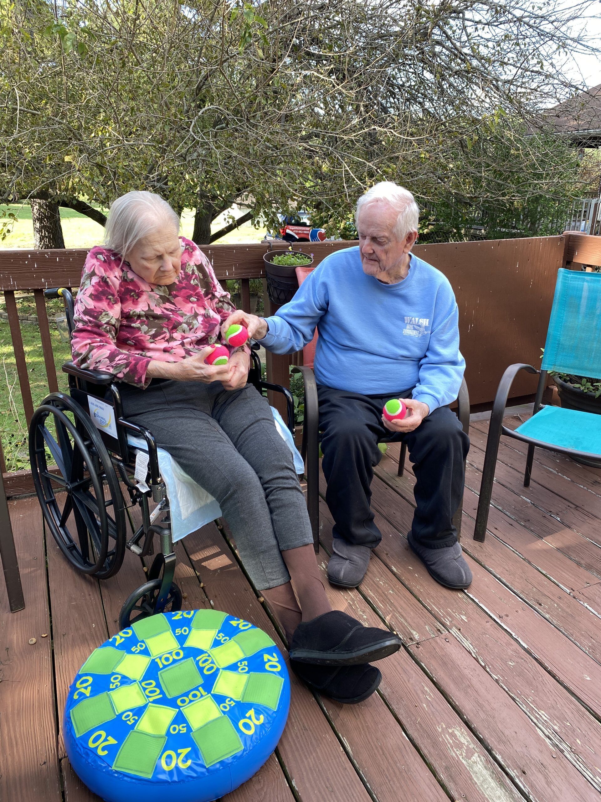 Two people in wheelchairs sitting on a wooden deck, sharing fruit with a colorful circular cushion nearby.