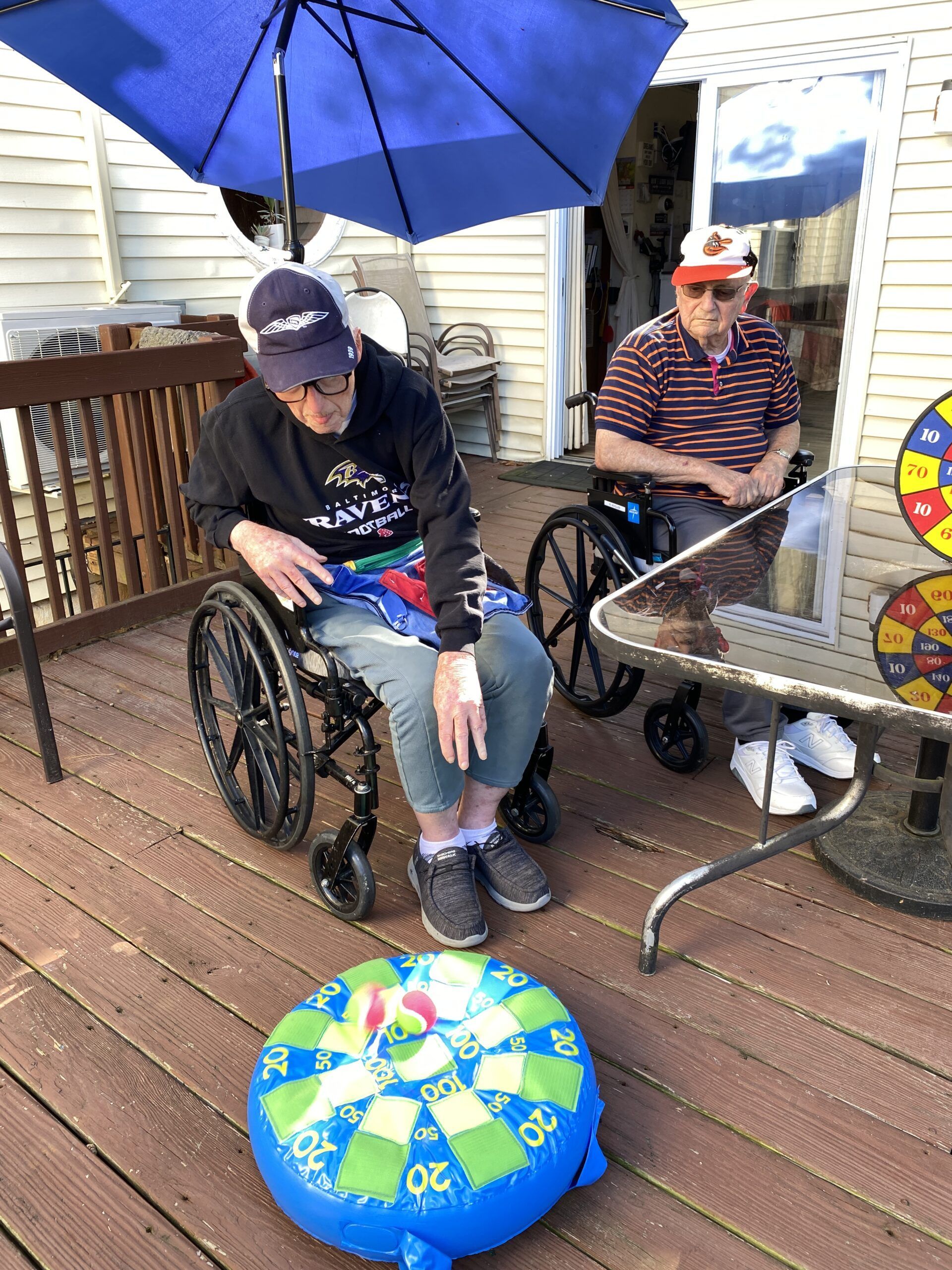 Two people in wheelchairs under a blue umbrella on a wooden deck, with a colorful beach ball in front.