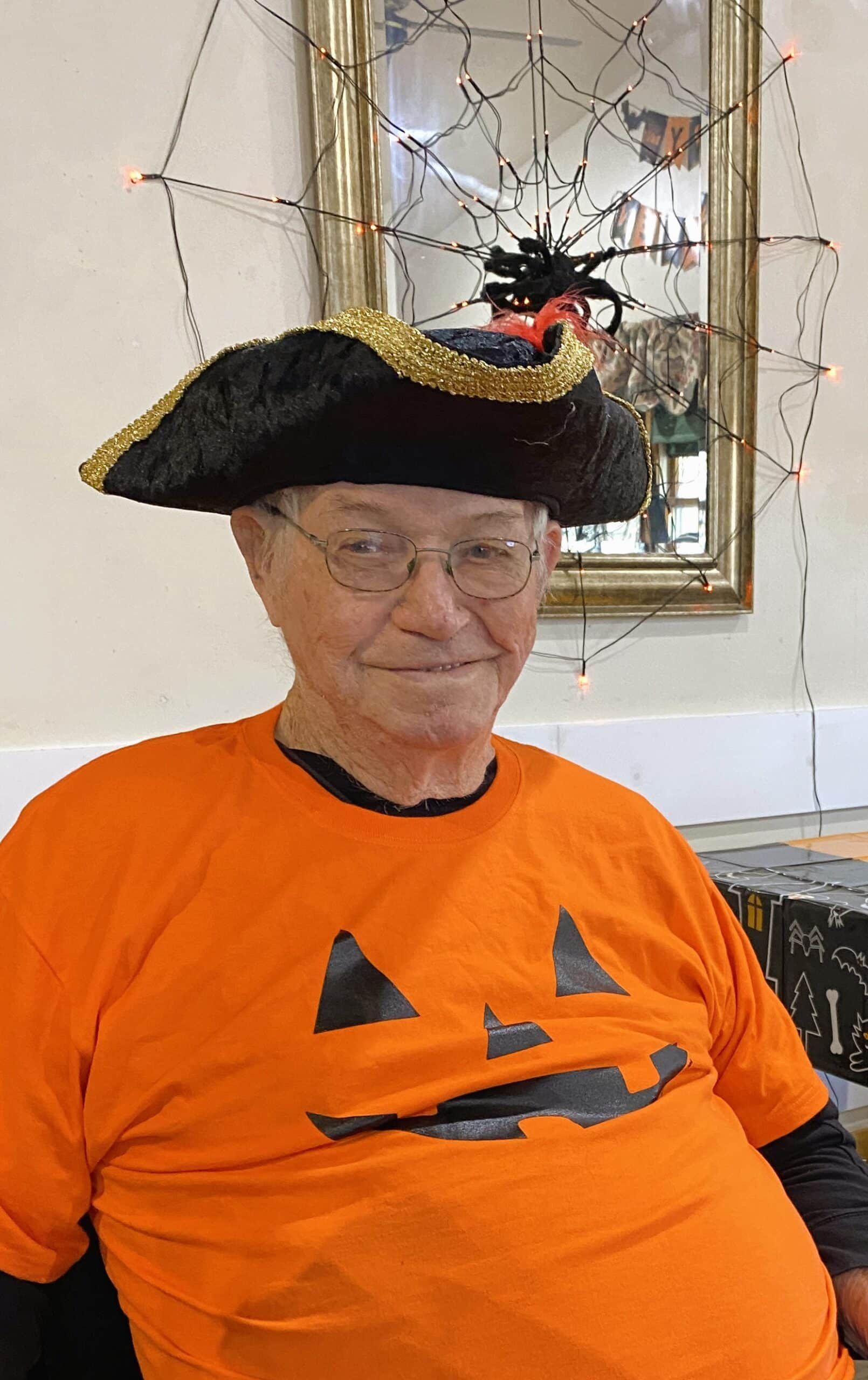 Person in an orange shirt and festive hat with a pumpkin face design, sitting indoors.