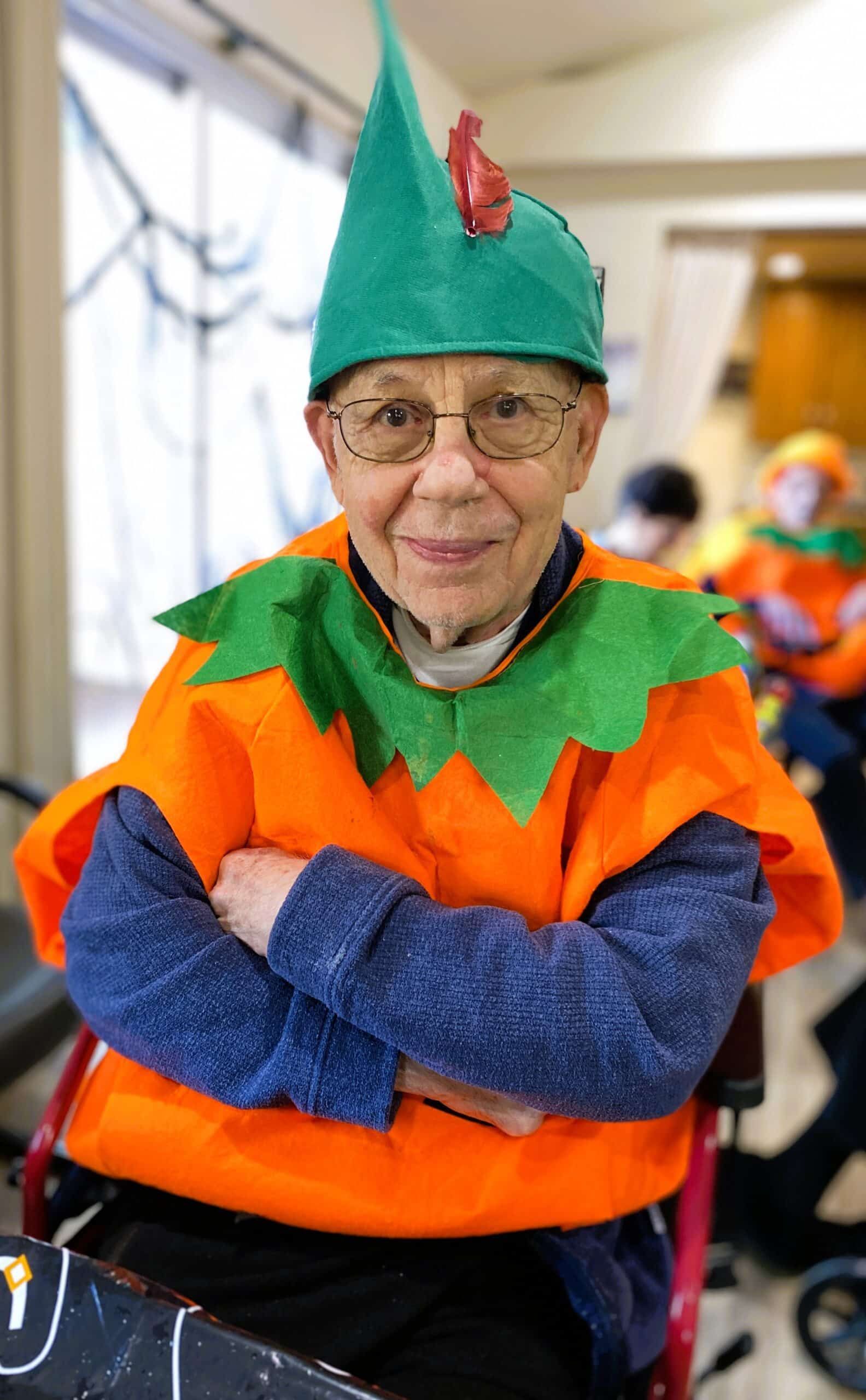 Person in a green leaf hat and orange costume, arms crossed, smiling indoors at a festive event