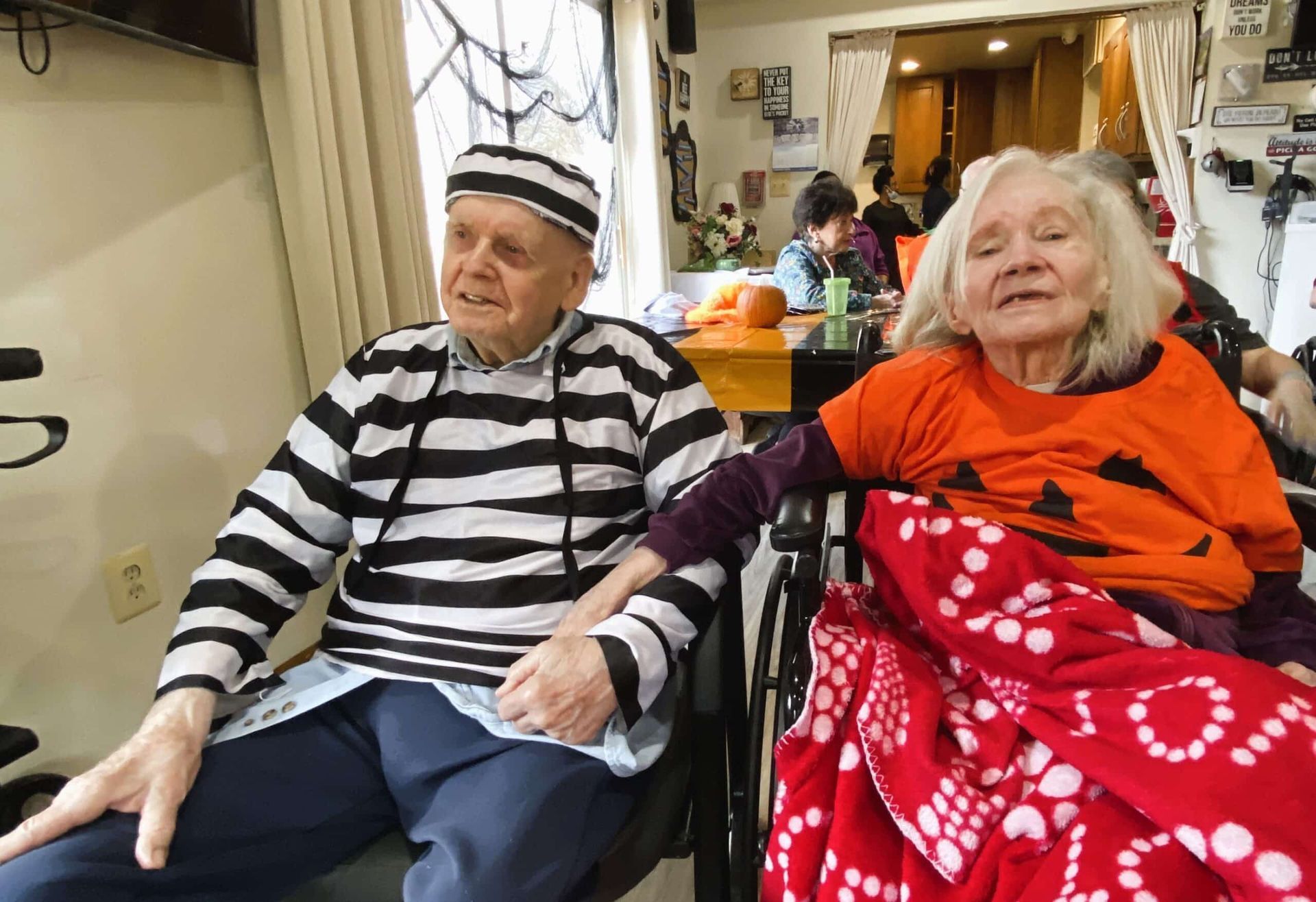 Two people seated indoors, wearing striped and orange clothing, with one holding a red polka-dot blanket