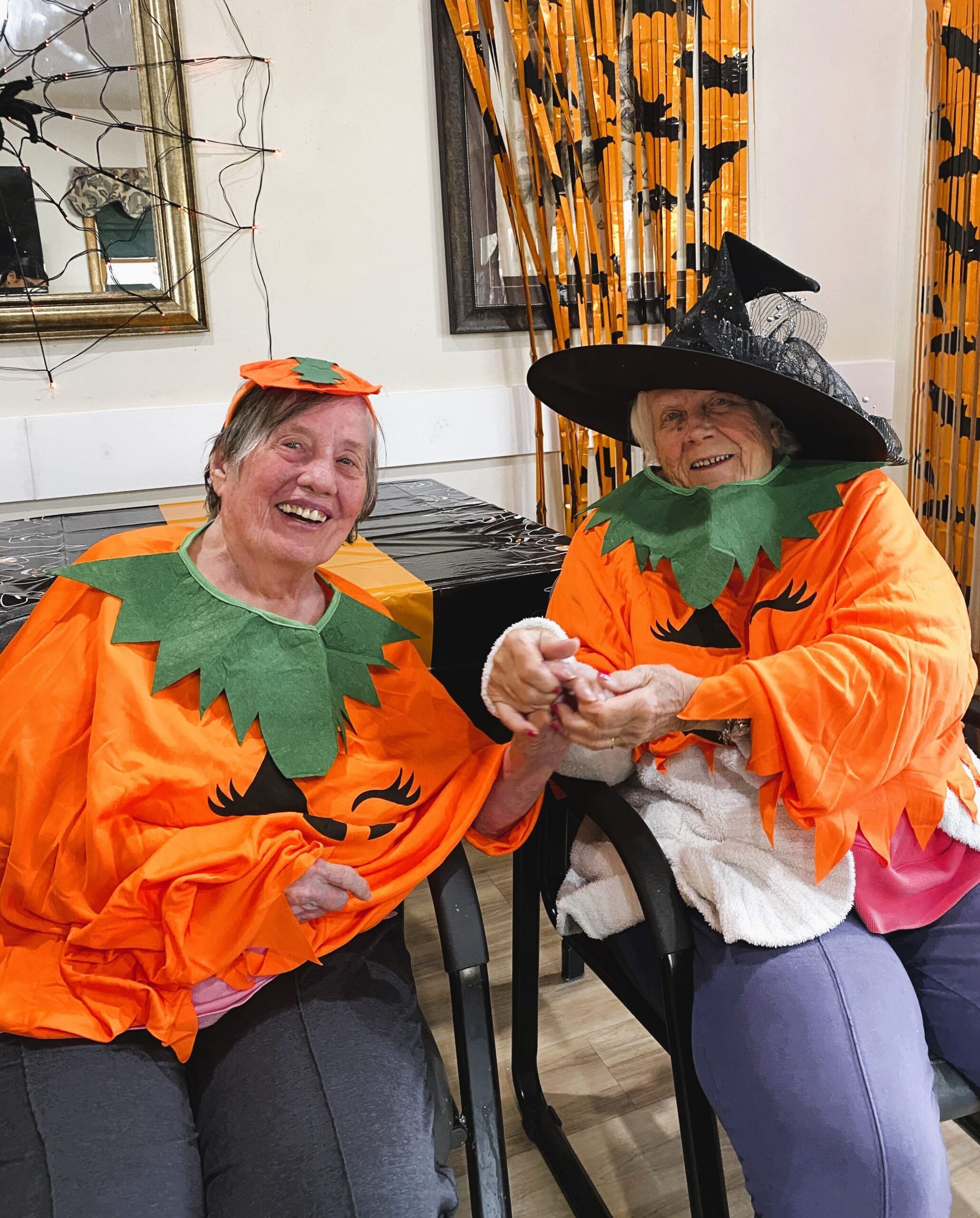 Two people in orange Halloween costumes and hats sit indoors, smiling and holding an object.