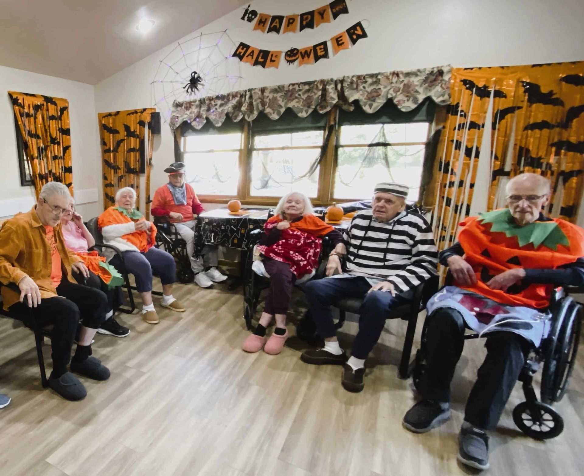 Senior adults seated in a decorated room, wearing Halloween costumes and hats, with orange-and-black bunting on the walls