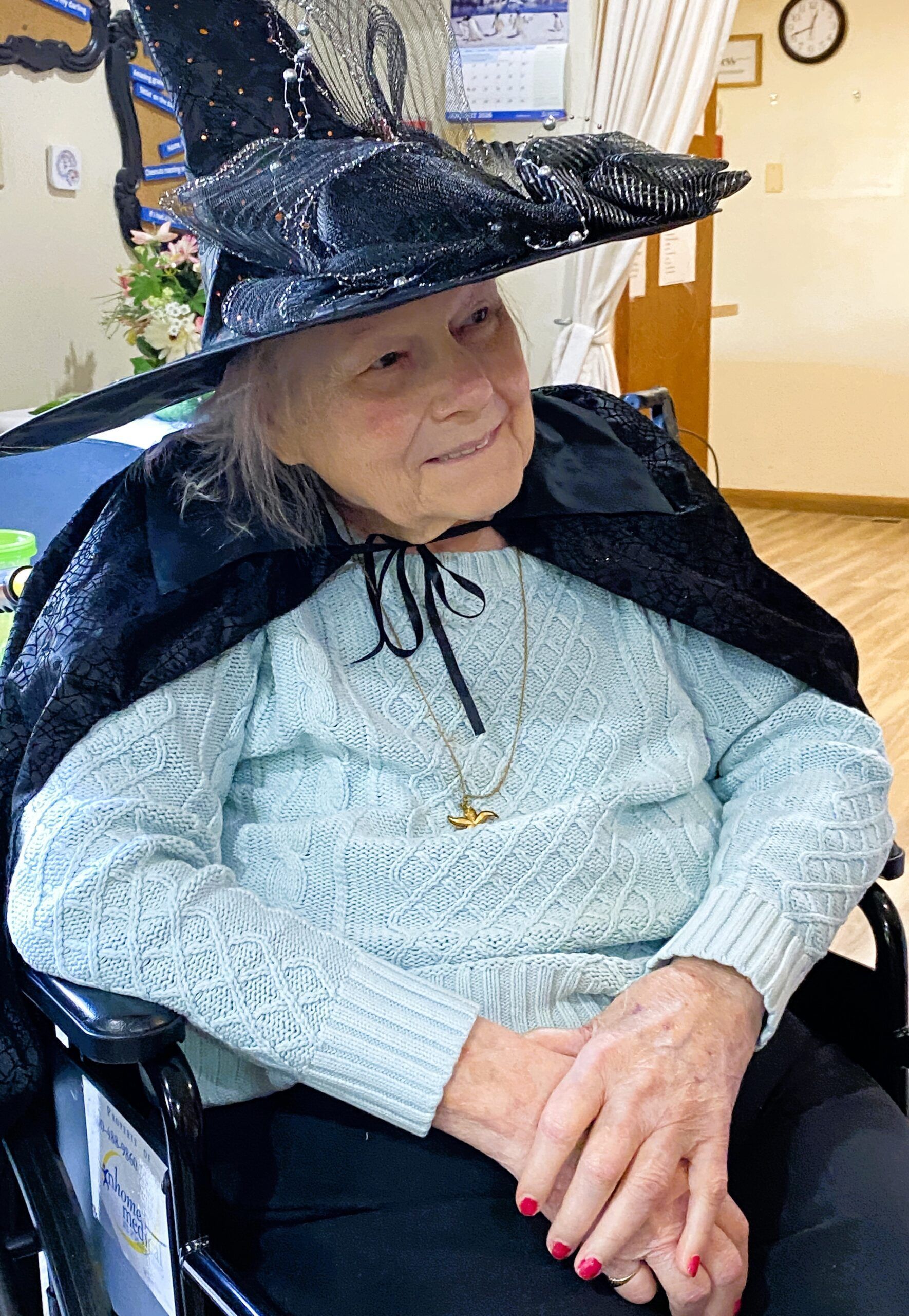 Person seated indoors wearing a large black hat and cape, with hands clasped and red-painted nails.