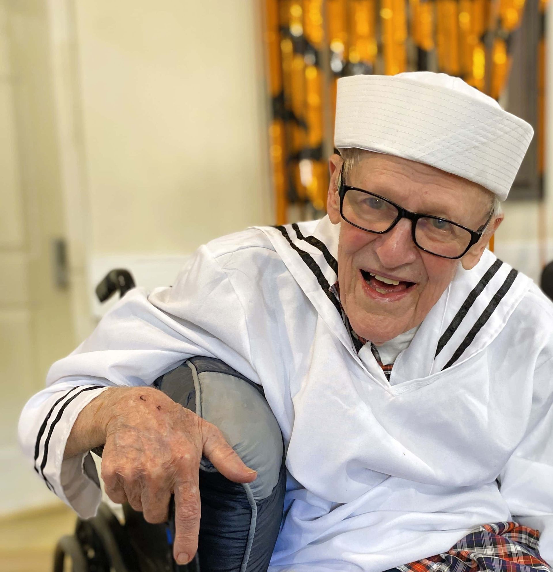 Smiling person in a white sailor outfit and hat, seated indoors with arm resting on a chair.